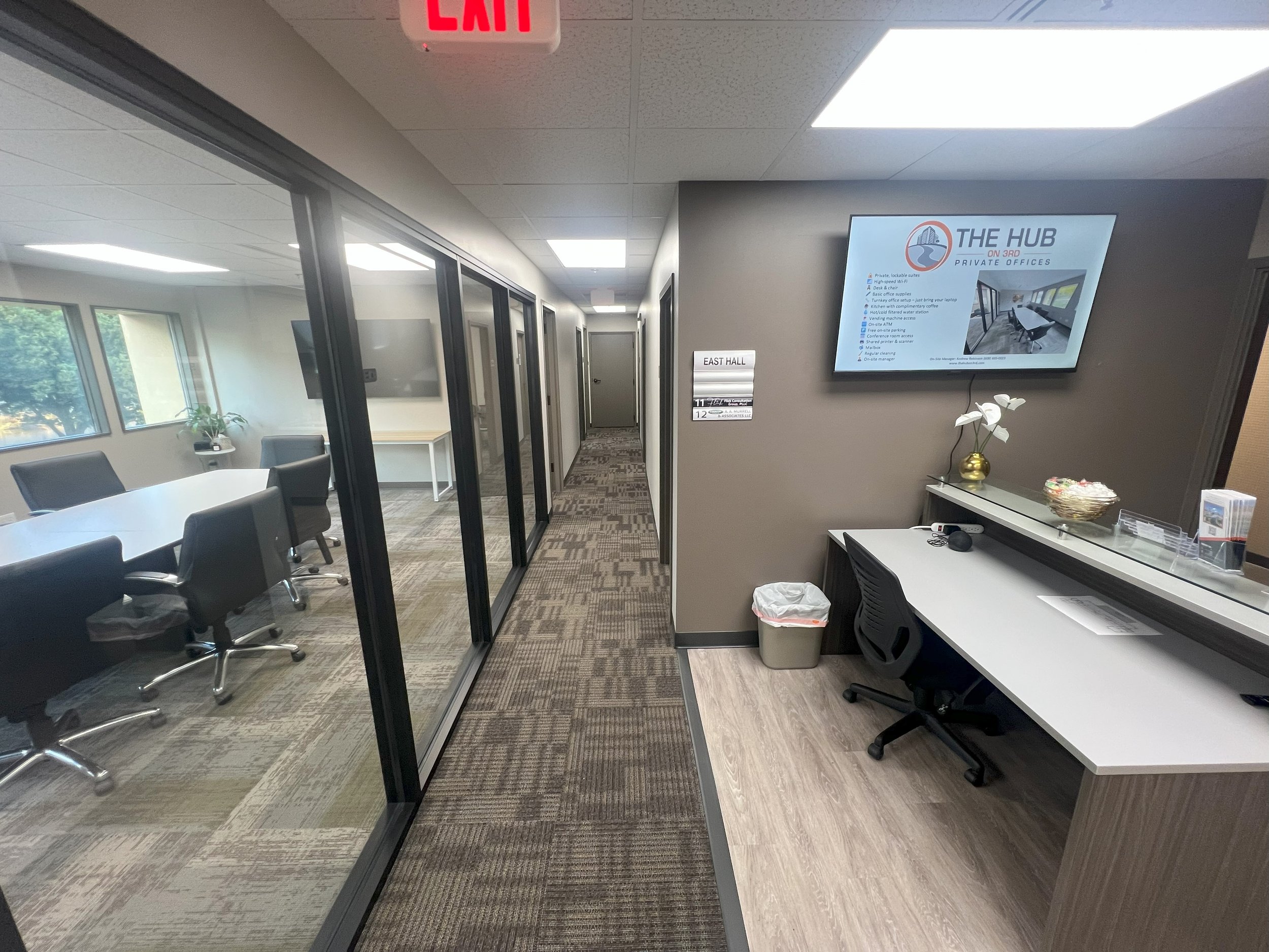 Interior view of an office space with a conference room on the left and a front desk with a computer on the right. There is a wall-mounted screen displaying a presentation titled "The Hub" on the right.