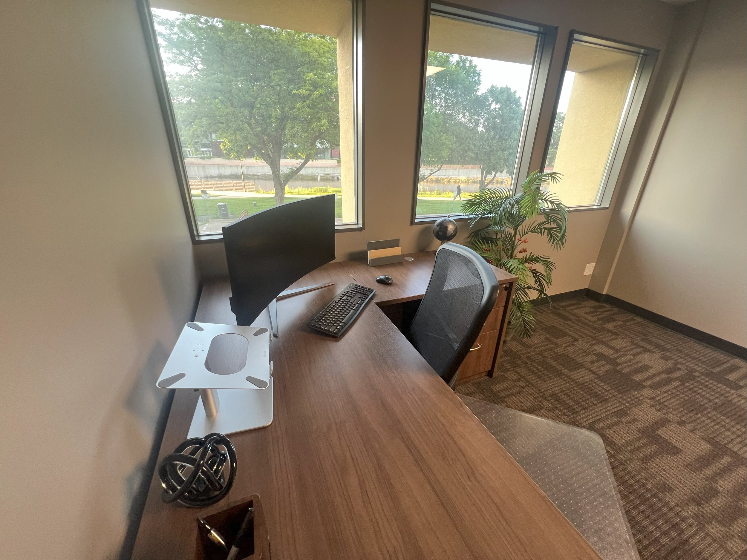 Office desk with a computer monitor, keyboard, mouse, desktop stand, and decorative globe near large windows with a view of trees and a park outside.