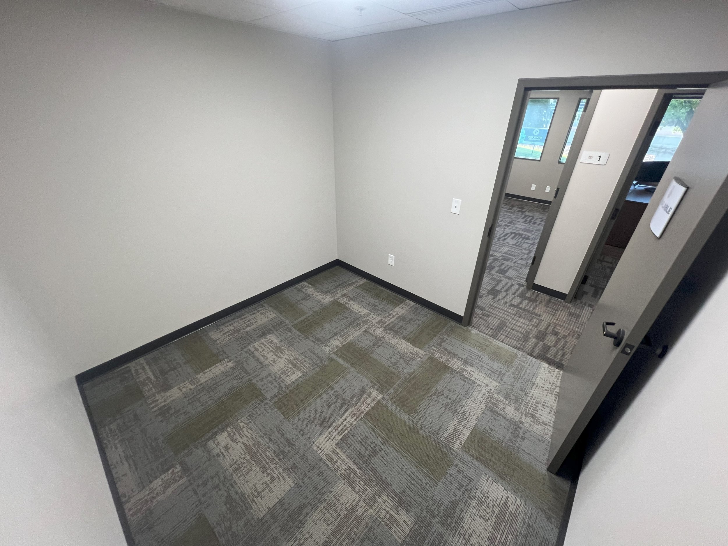 Empty office room with beige walls, patterned carpet, and an open doorway leading to another room with similar decor and a window.