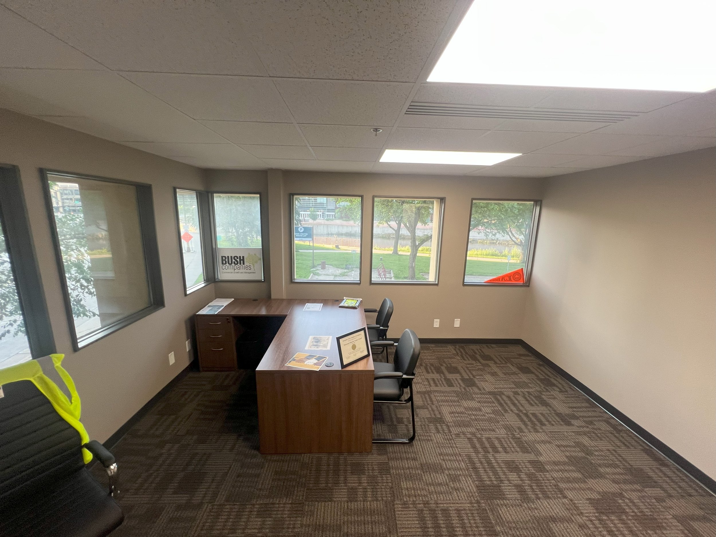 An empty office with a wooden desk, two chairs, windows showing an outdoor view, and various papers and a framed certificate on the desk.