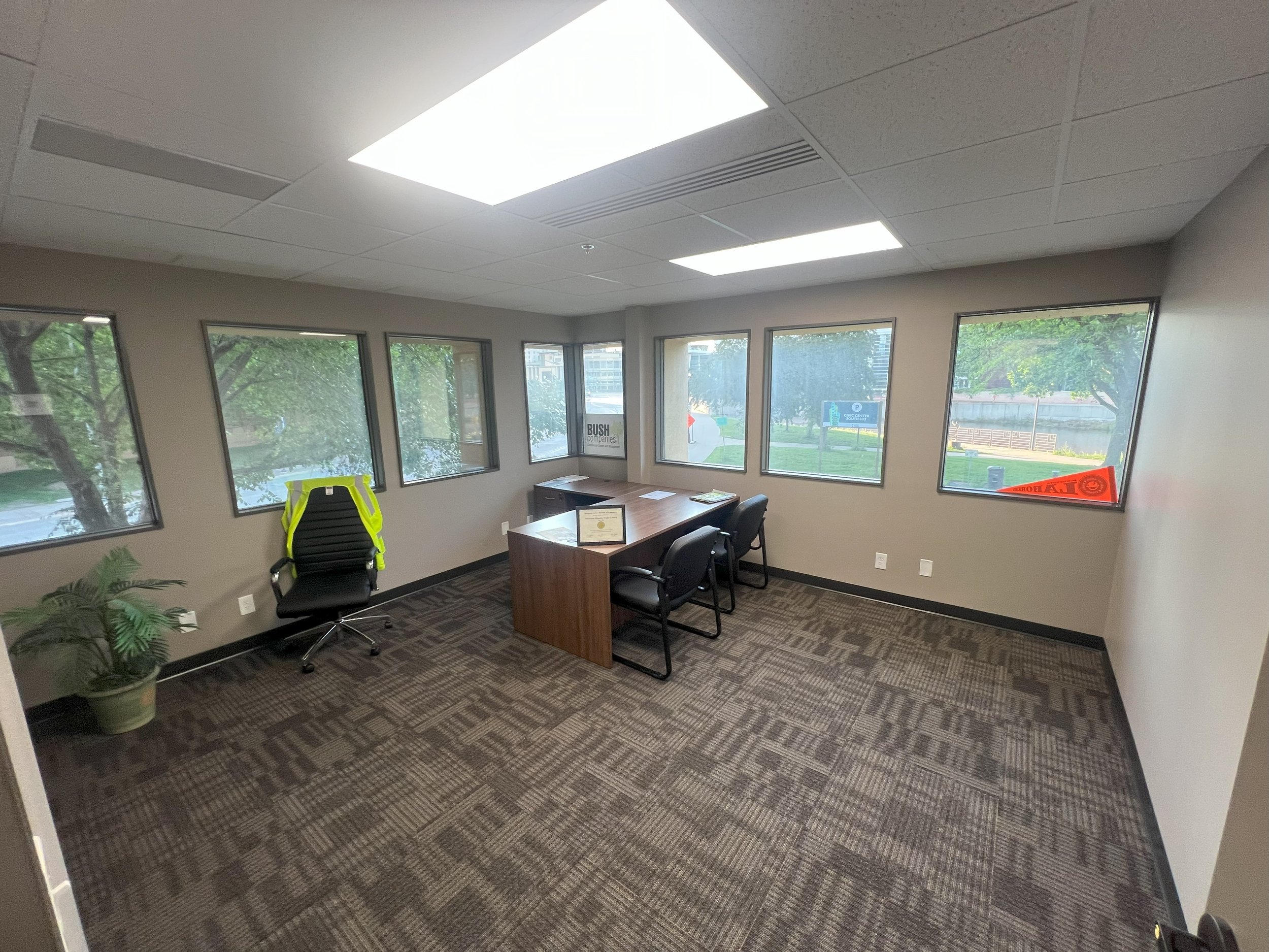 Empty office with a desk, three chairs, a plant, and a high-visibility vest on a chair. Large windows showing trees outside.