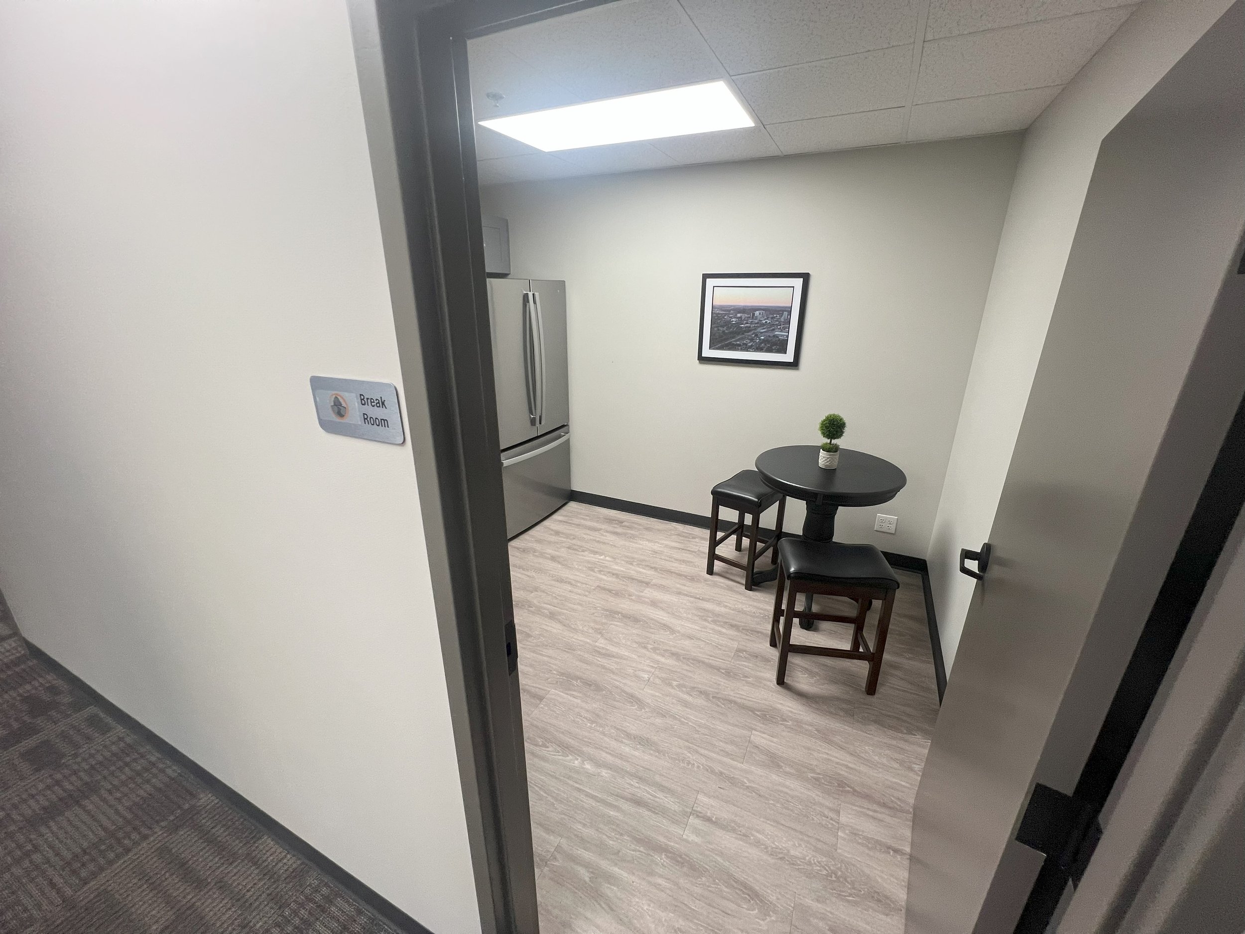 Small break room with a stainless steel refrigerator, a framed picture on the wall, a round black table with a small potted plant, and two black stools. Sign on the wall reads 'Break Room'.