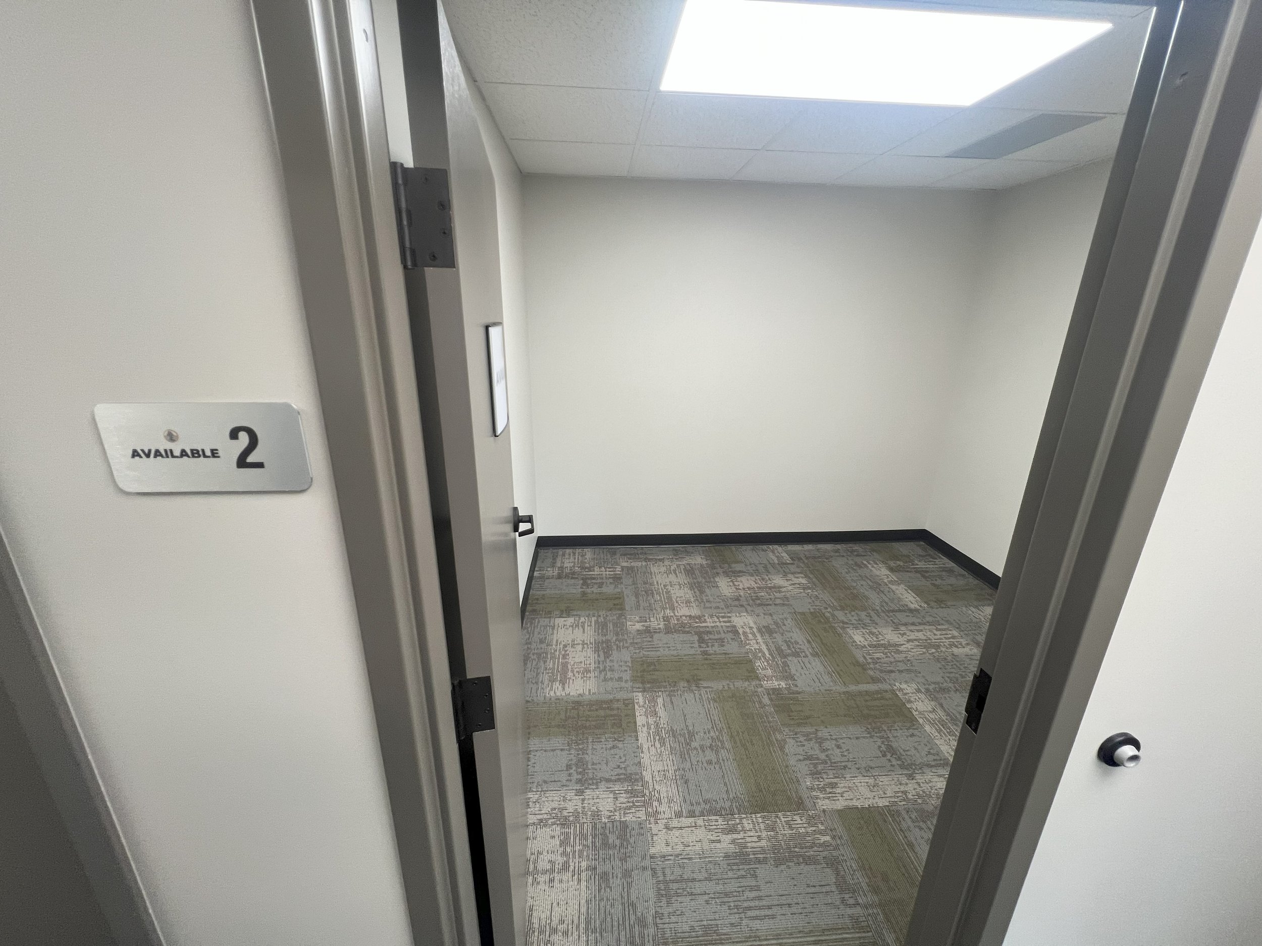 Empty office or storage room with beige walls, patterned carpet, a door slightly open, and a sign reading 'Available 2' outside.