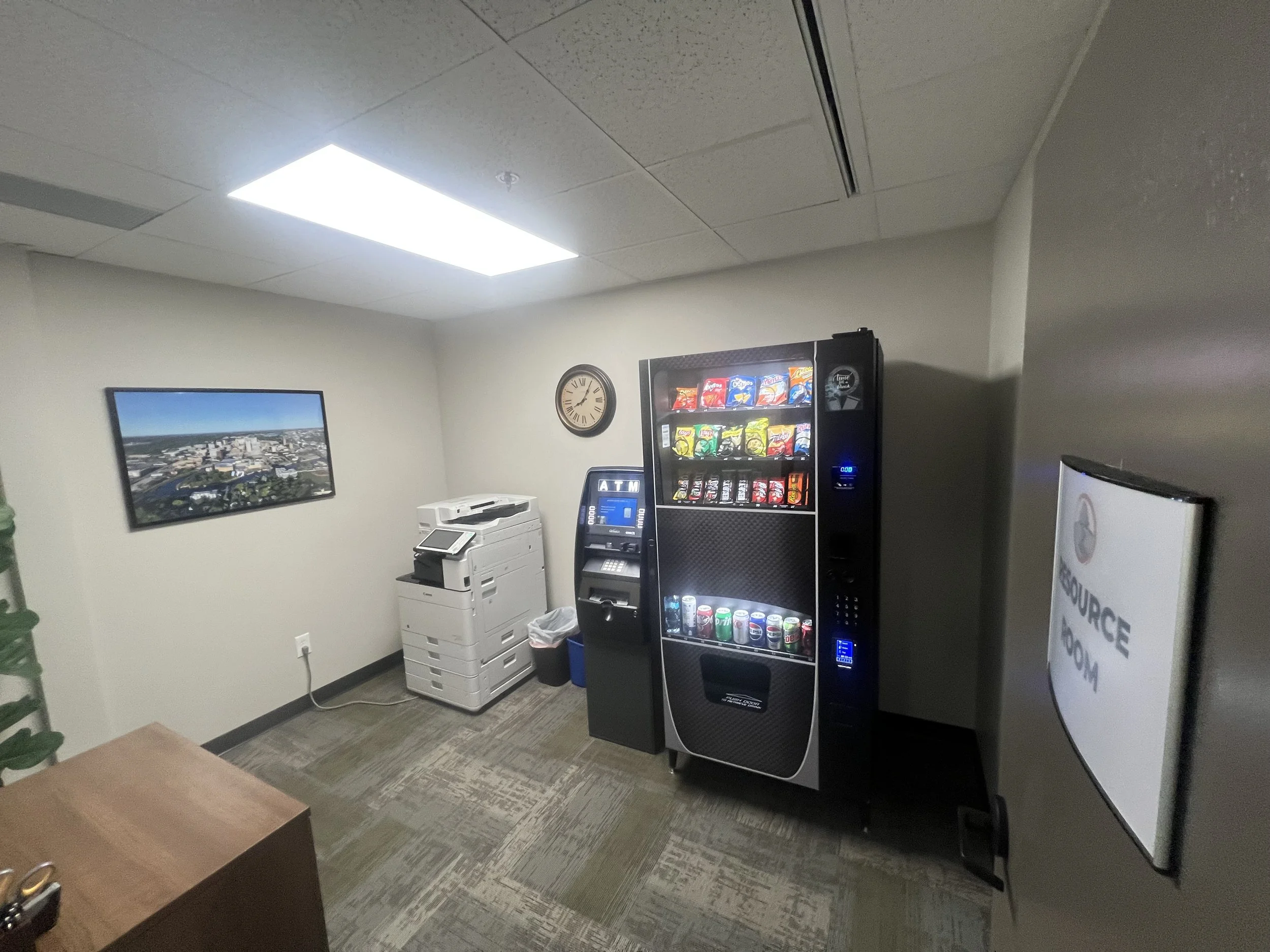 Small office room with vending machine, ATM, photocopy machine, wall clock, and framed aerial cityscape photo.