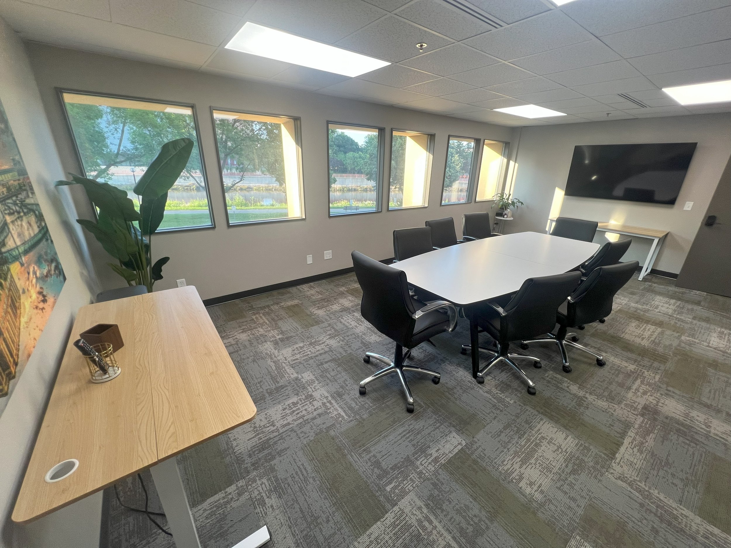 Empty conference room with large window, seven black chairs around a white oval table, a wall-mounted TV, a plant, and a wooden side table with office supplies.
