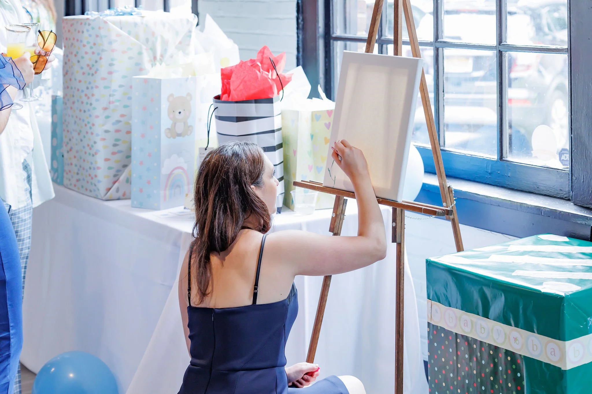 A woman painting on a canvas at a baby shower decorated with gift bags, balloons, and a table of presents near a large window.