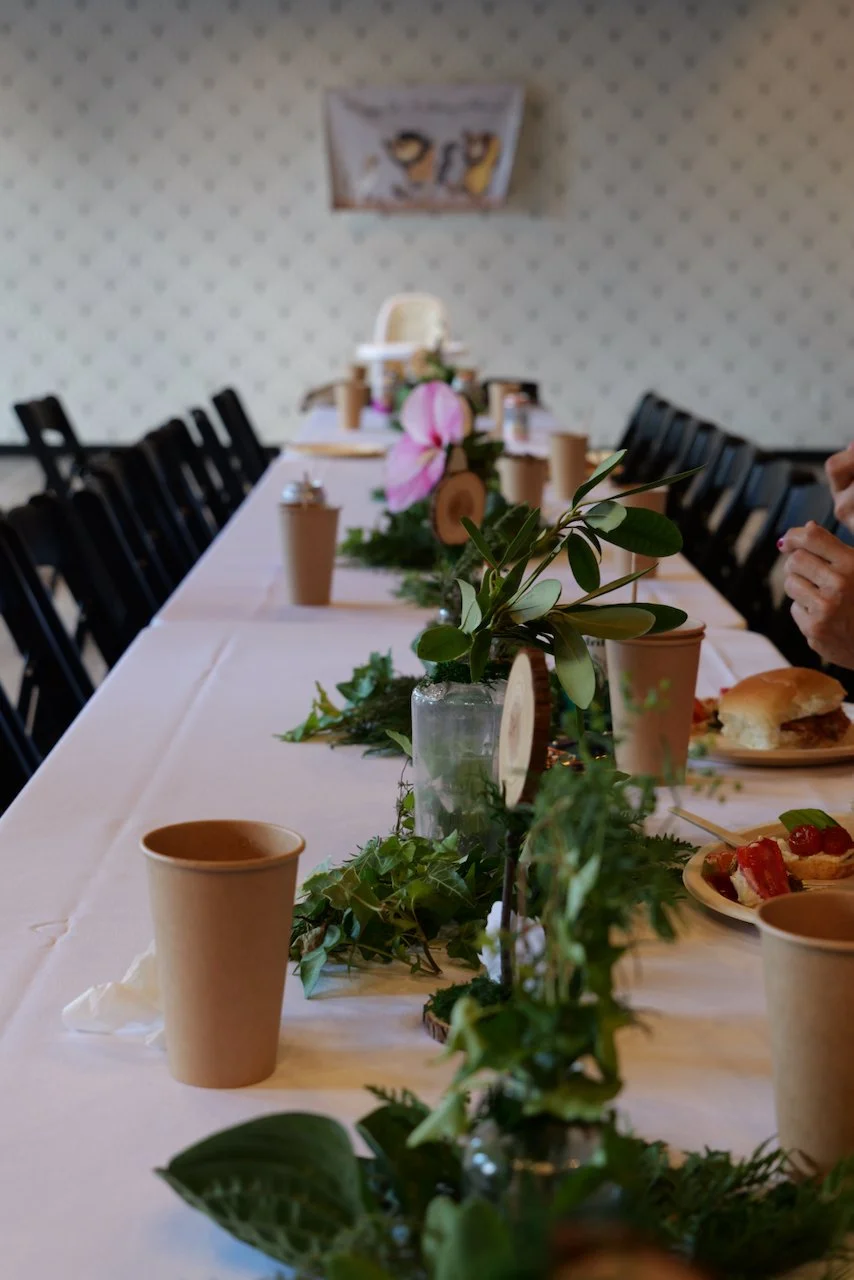 Long dining table decorated with a white tablecloth, greenery, and pink flower centerpieces, set for a meal with paper cups, plates of food, and a few hands visible.
