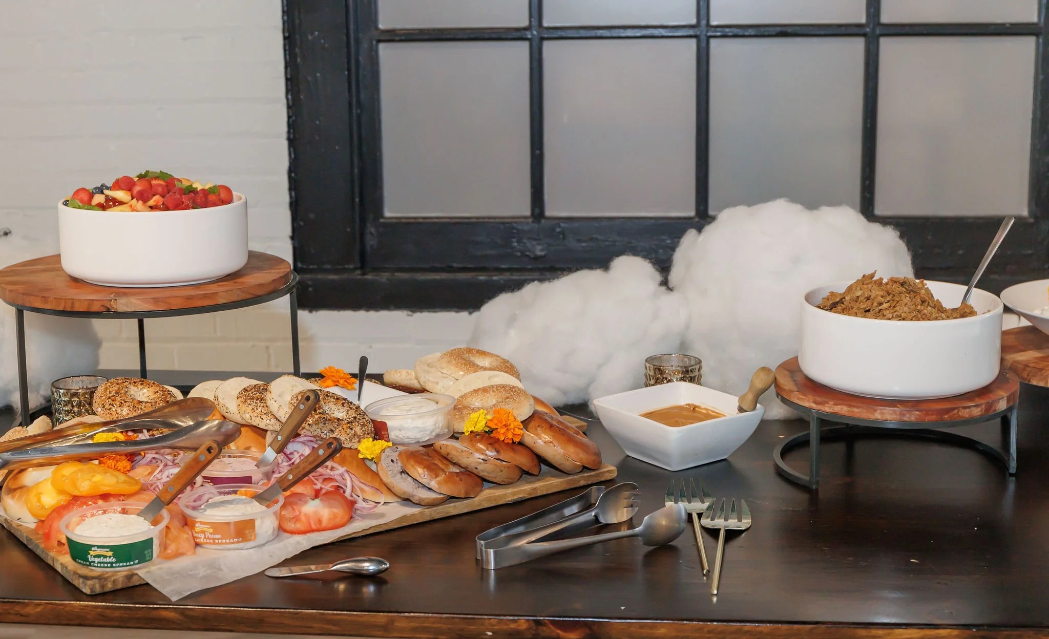 Assorted breakfast foods including bagels, bread rolls, sliced tomatoes, sliced onions, sliced meats, cheeses, spreads, and a large bowl of cereal, arranged on wooden and elevated surfaces with decorative flowers and eating utensils.