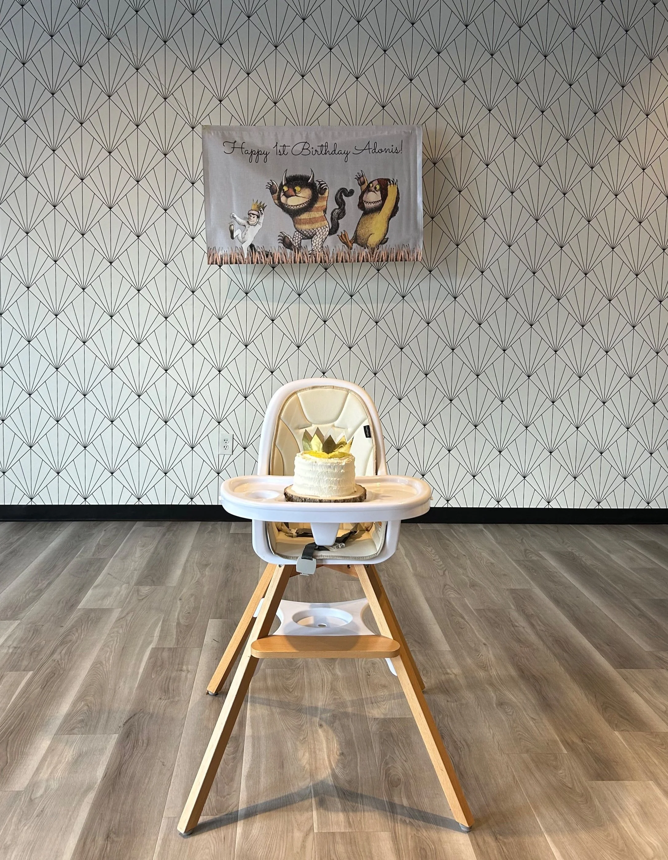 A birthday cake with a crown decoration on top, placed in a high chair in a room with geometric patterned wallpaper. There is a birthday banner with characters on the wall that reads 'Happy 1st Birthday Adonis!'