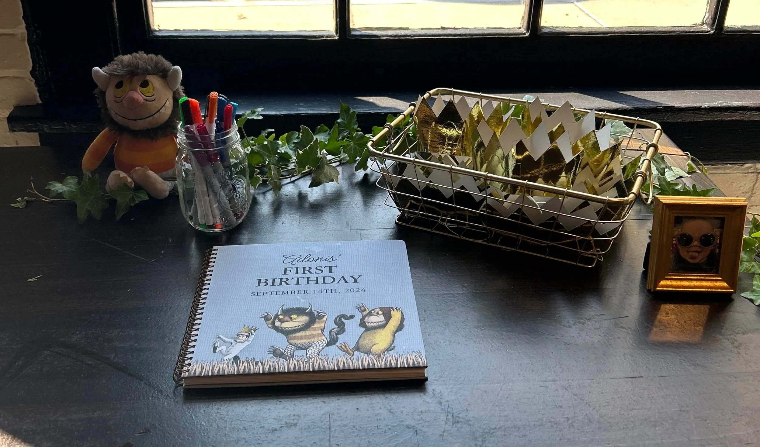 Table decorated for a child's first birthday with a first birthday book, toy doll, picture frame, pen jar, gold and white paper decorations, and ivy plants next to a window.
