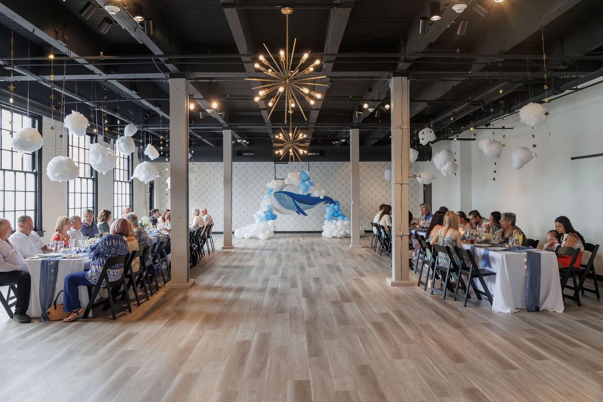 A decorated event space with long dining tables, white tablecloths, and hanging cloud-like decorations, featuring a blue whale balloon centerpiece, for a celebration or party.