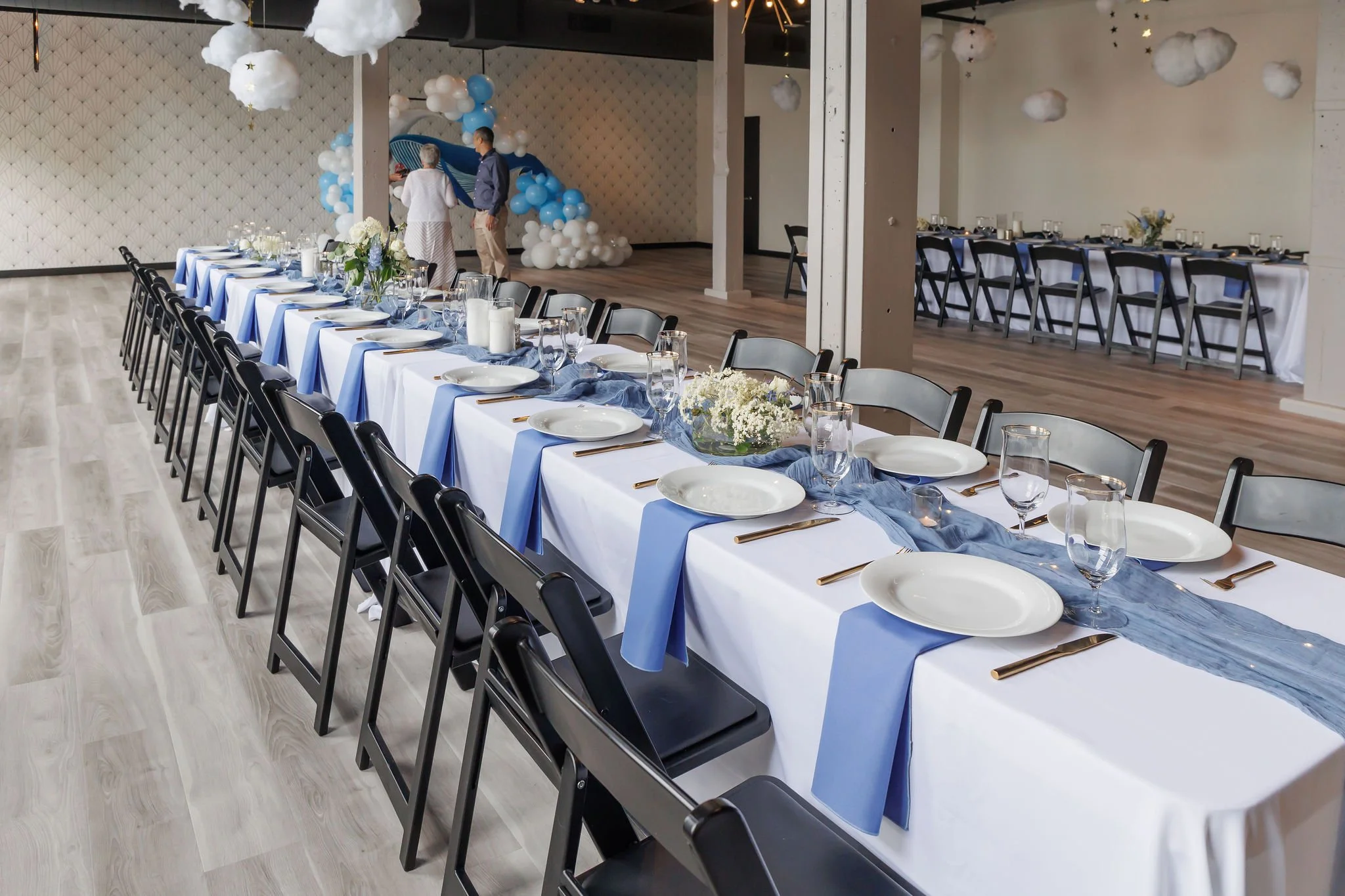 Long banquet table with white tablecloth, blue cloth runners, white plates, gold utensils, and stemware, in a decorated event hall with white and blue balloon arch and cloud-shaped hanging decorations.