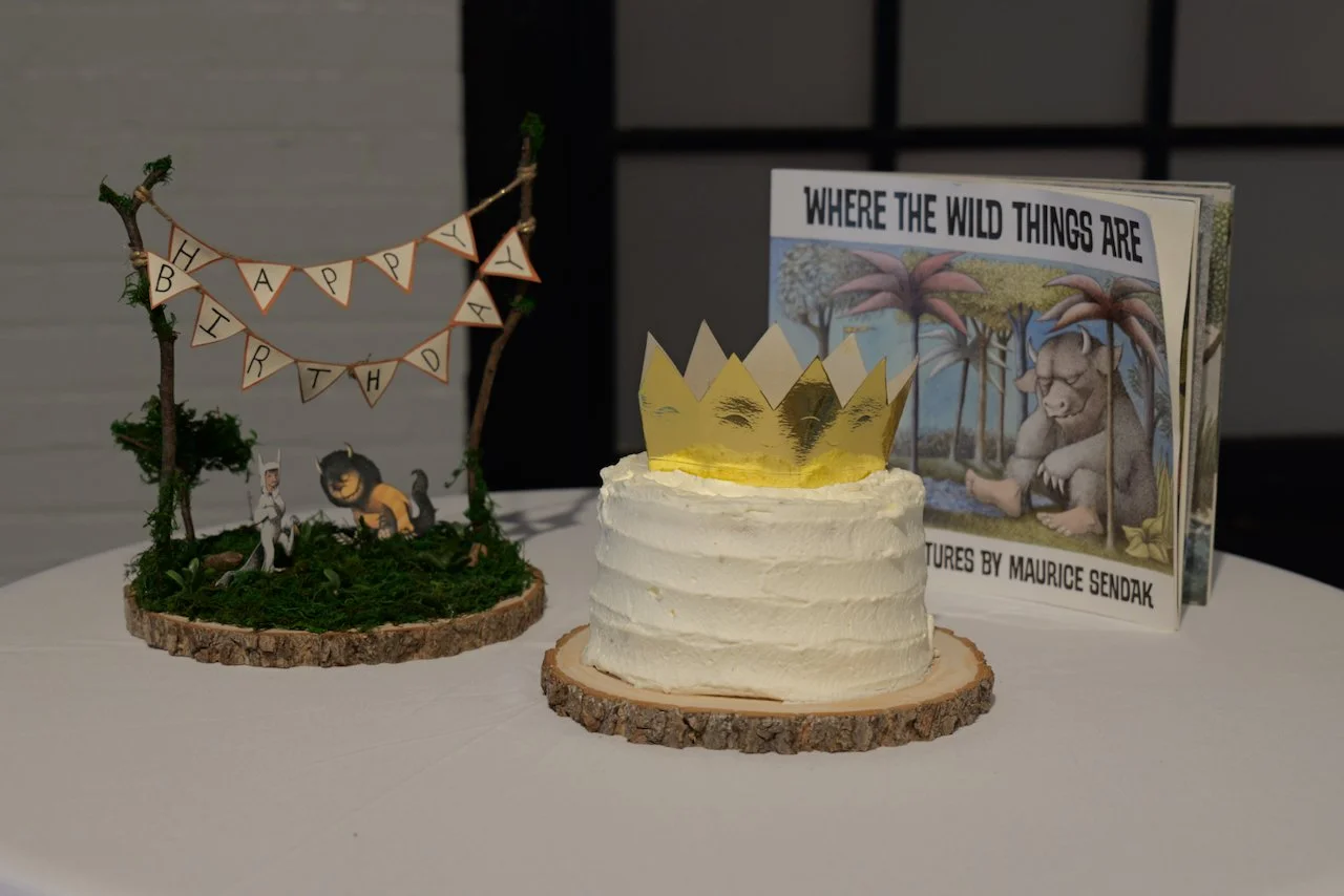 A birthday cake with a gold paper crown on top, next to a book titled "Where the Wild Things Are" and a decorative sign with a "Happy Birthday" message, all on a white table.