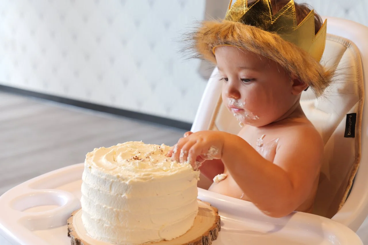 A young child wearing a gold crown and a furry gold hat, sitting in a high chair and reaching for a birthday cake with white frosting, on a wooden cake board.