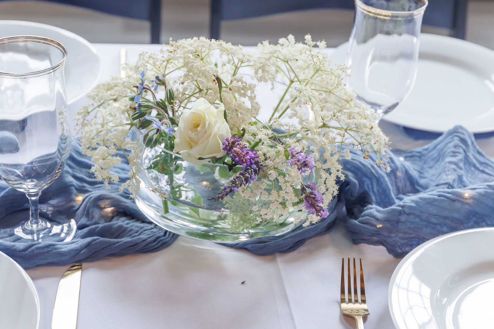 Elegant table setting with a floral centerpiece of white and purple flowers, blue table runner, wine glasses, and white dishware.