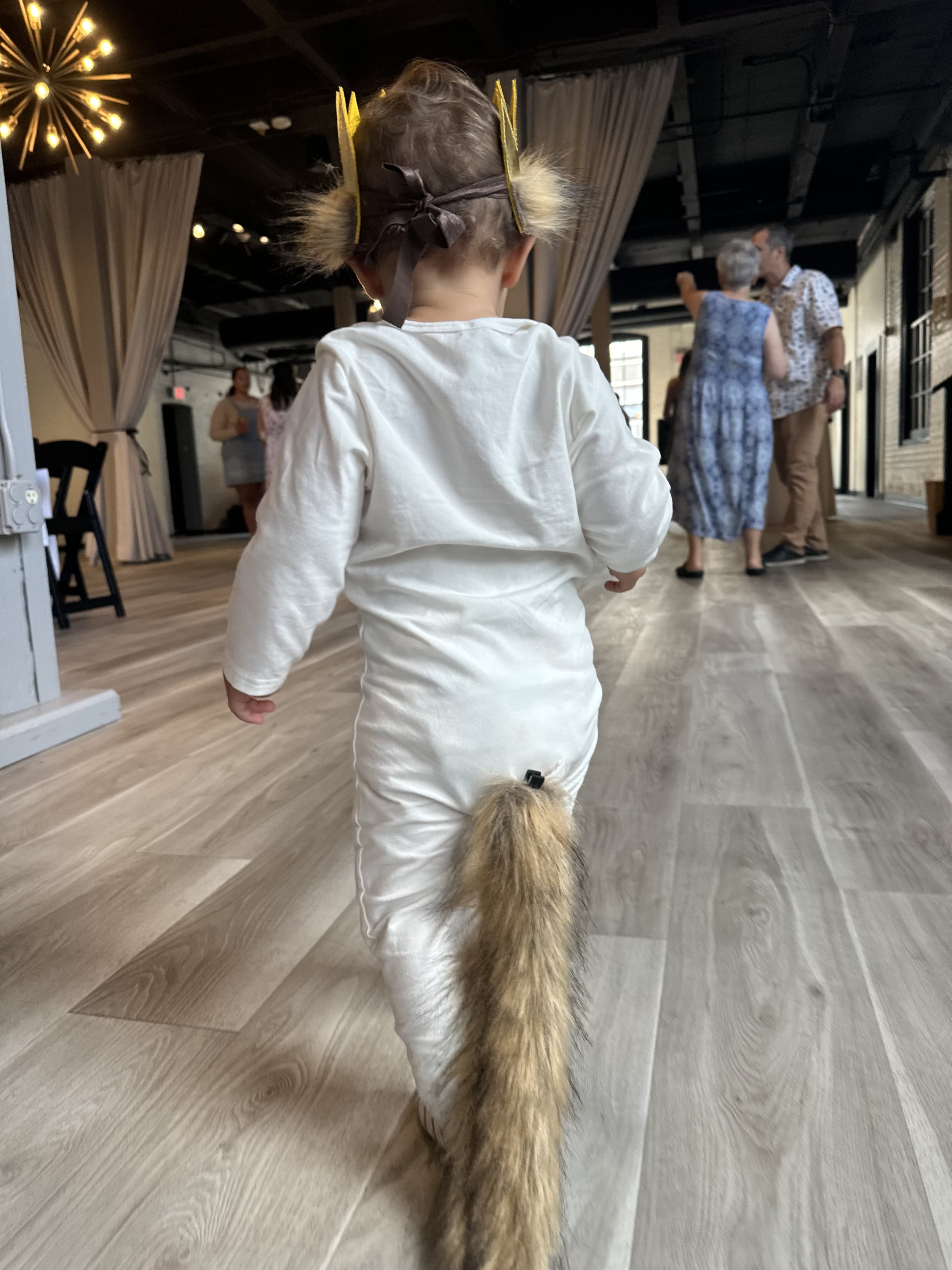 Child dressed in a fox costume with a tail, walking indoors during a gathering.