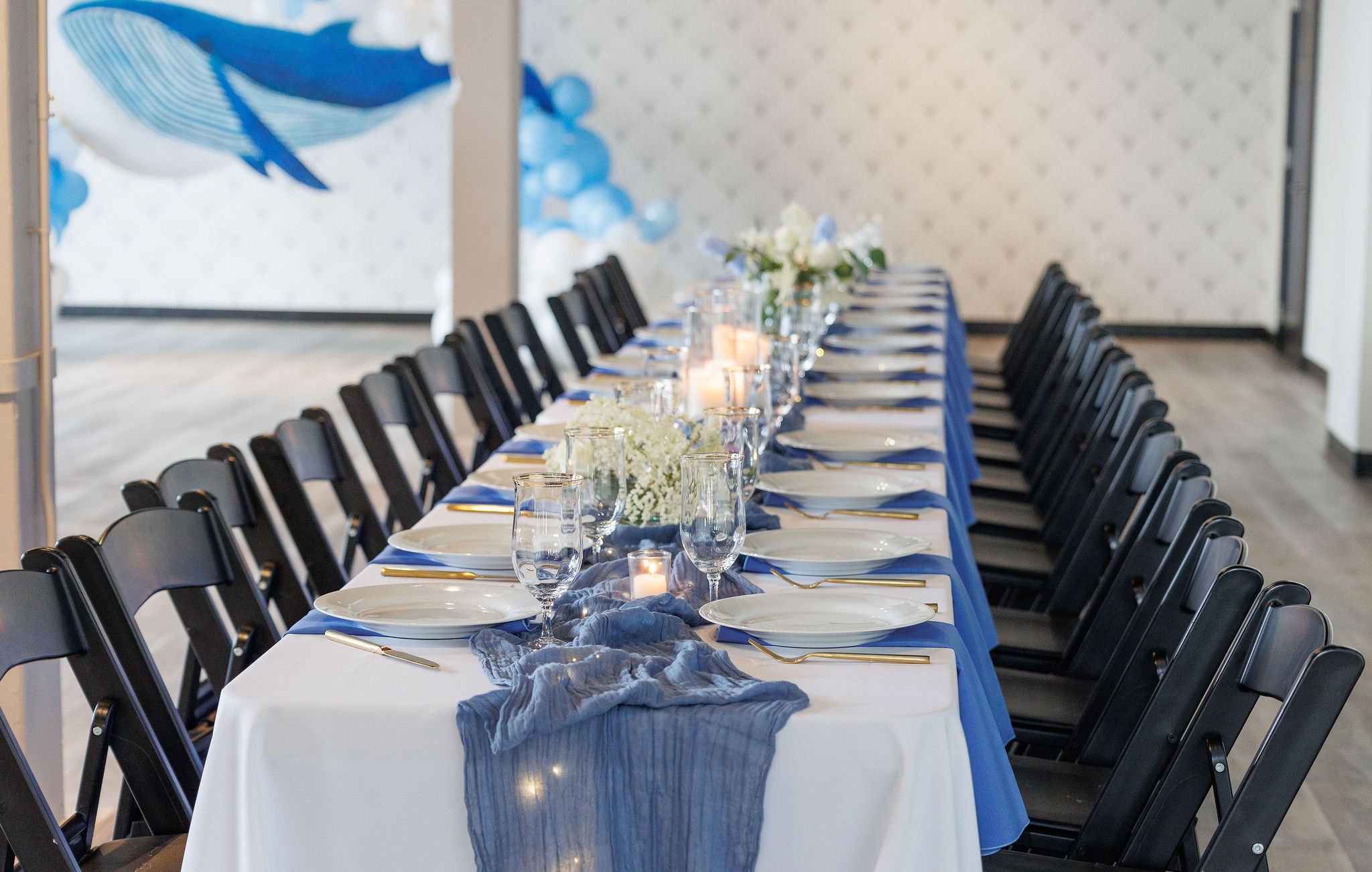 Long banquet table decorated with white tablecloth, blue runner, floral centerpieces, candles, and set with white plates, gold utensils, and drinking glasses, arranged for a formal event or celebration.
