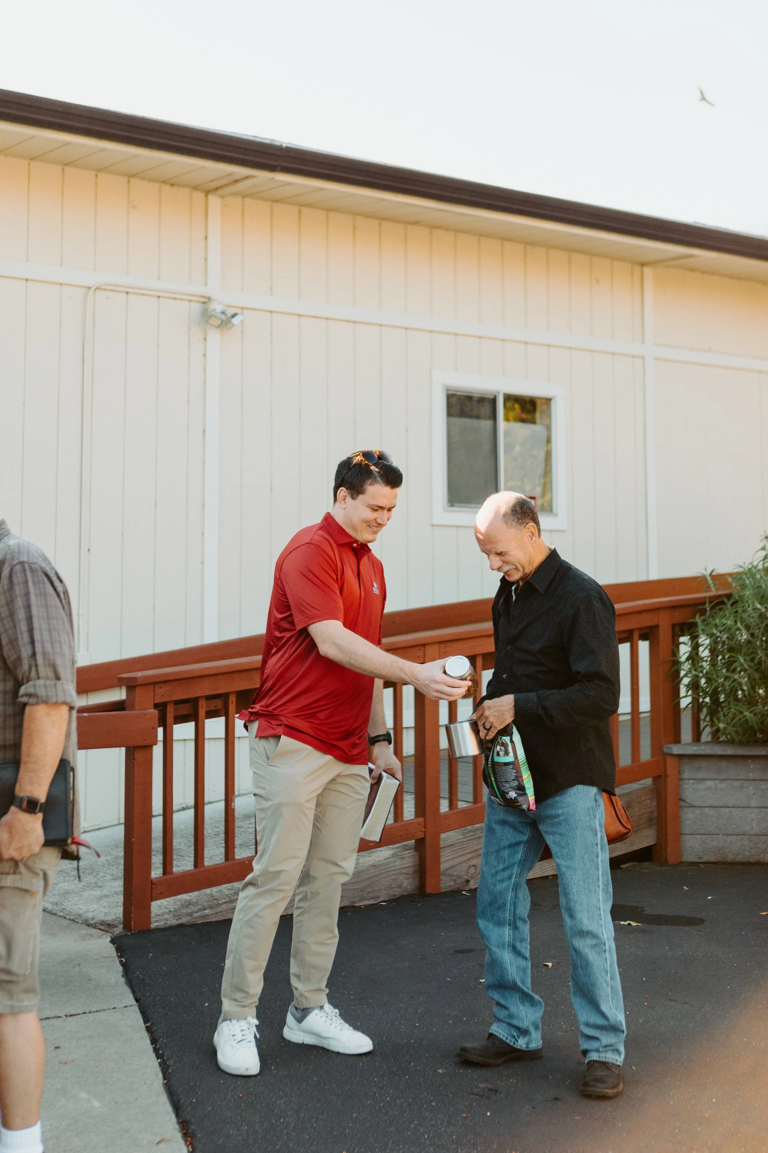 Two men smiling, one handing a small container to the other outside a building with a wooden railing.