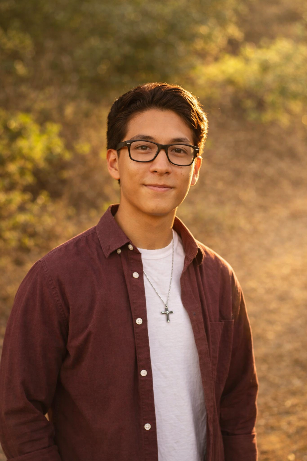 A young man with short dark hair, glasses, wearing a white t-shirt, a maroon button-up shirt, a cross necklace, and a watch, standing outdoors in front of a gray corrugated metal wall.