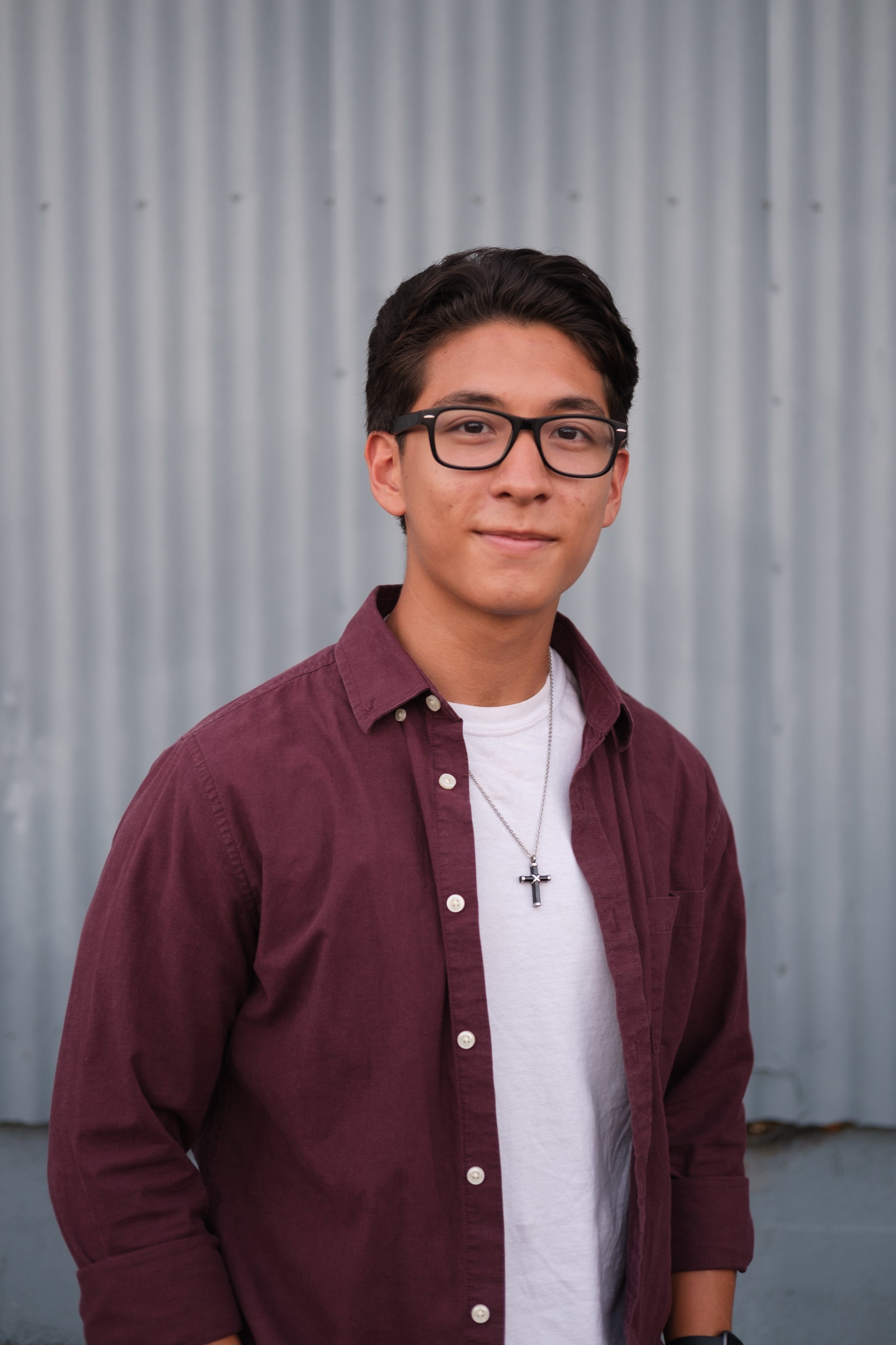 A young man with short dark hair, glasses, wearing a white t-shirt, a maroon button-up shirt, a cross necklace, and a watch, standing outdoors in front of a gray corrugated metal wall.