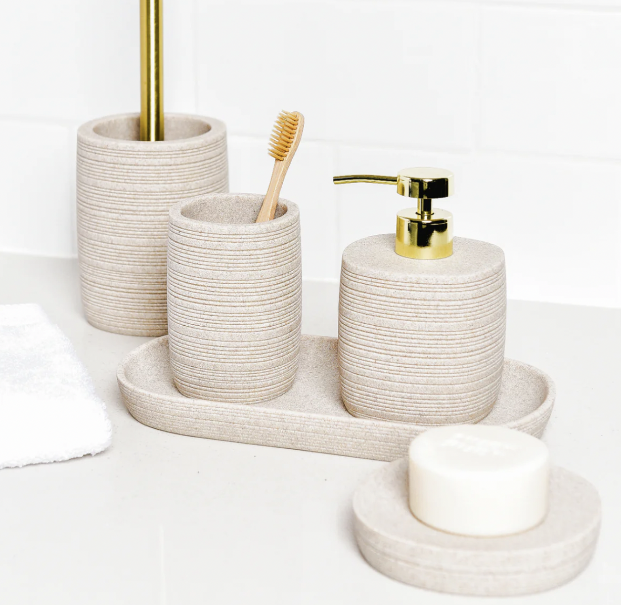 Bathroom accessories including beige textured ceramic holders, a toothbrush, a soap dispenser with a gold pump, a bar of soap on a ceramic dish