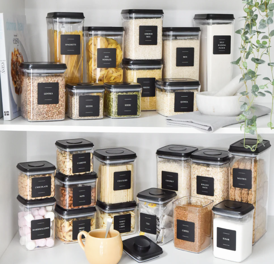 Pantry shelf with labeled food storage jars containing pasta, rice, flour, grains, seeds, nuts, cereal, sugar, coffee, and other dry foods, with a mortar and pestle and a potted plant nearby.