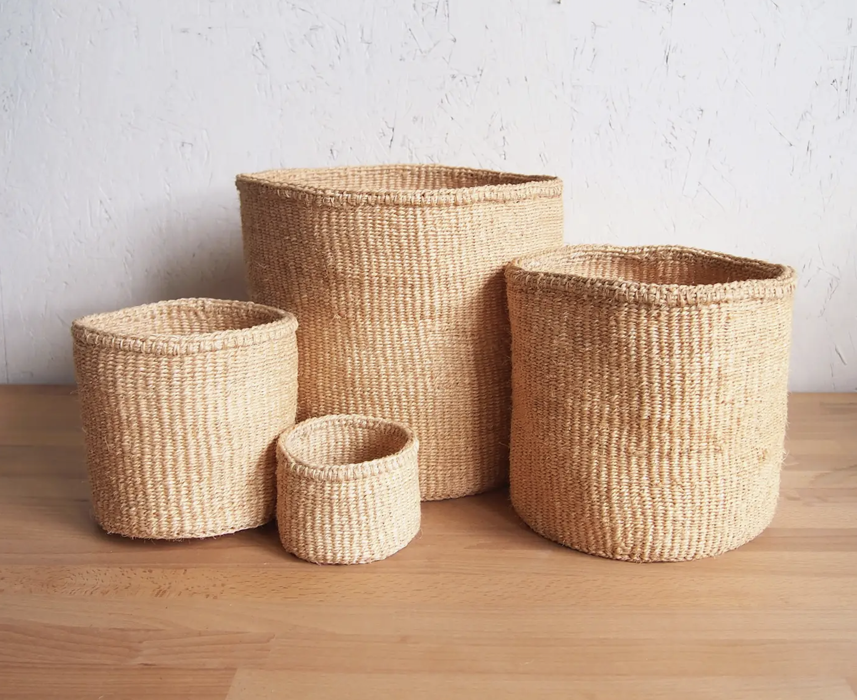 Set of five woven storage baskets in natural color, varying in height, arranged on a wooden surface against a white textured wall.