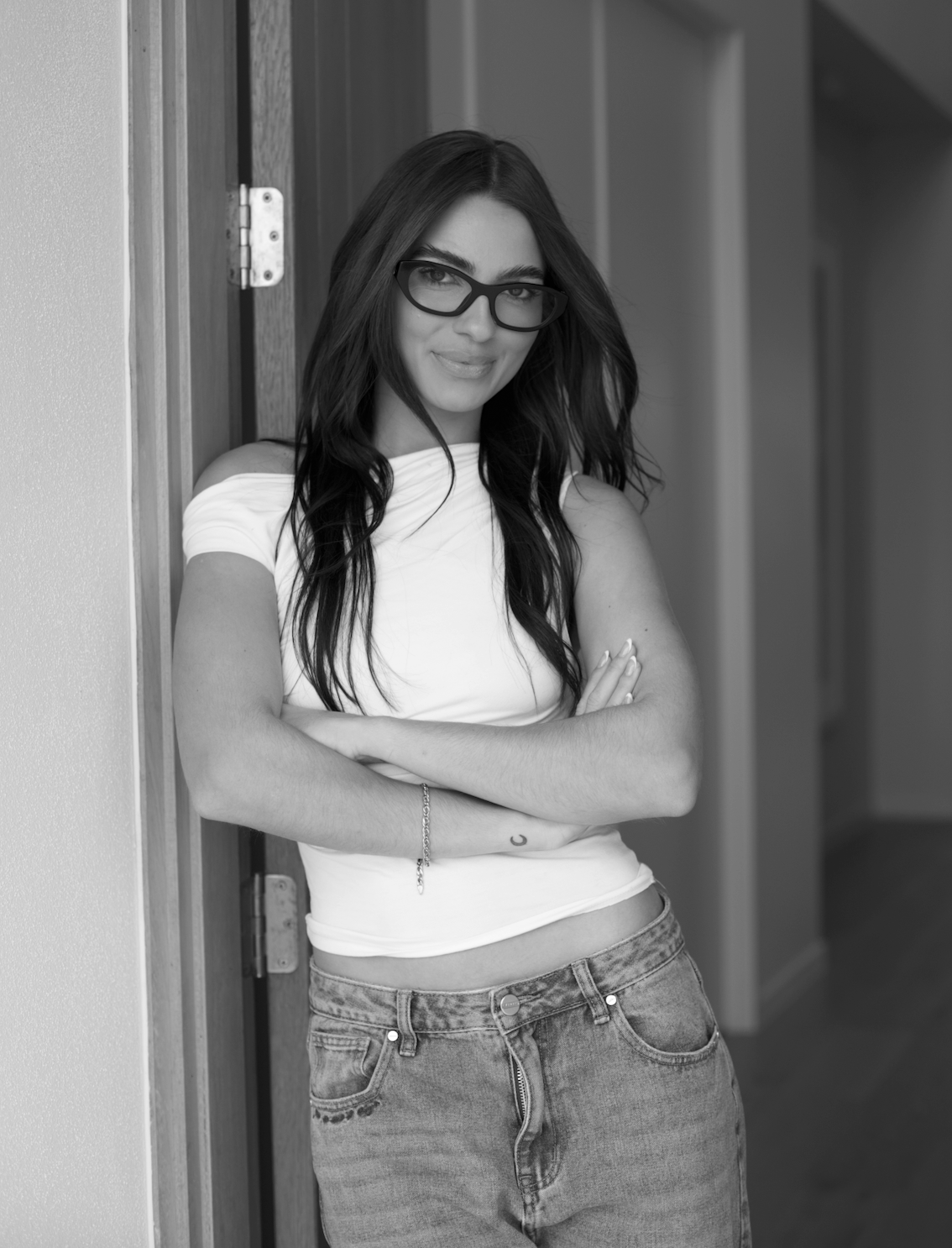 Black and white photo of a young woman with long dark hair, wearing glasses, a white off-shoulder top, and jeans, leaning against a wall with arms crossed and smiling.