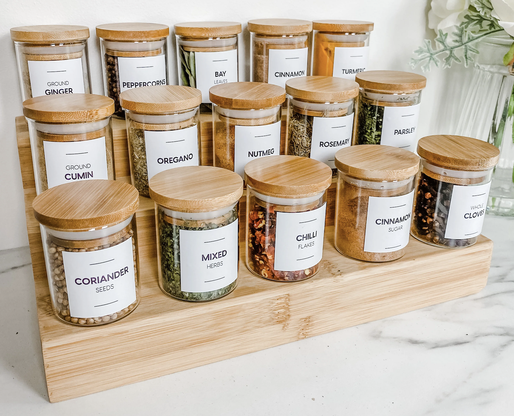 A wooden spice rack holds glass jars with wooden lids containing various spices and herbs labeled white labels with black text, including ground ginger, peppercorns, bay leaves, cinnamon, turmeric, ground cumin, oregano, nutmeg, rosemary, parsley, co