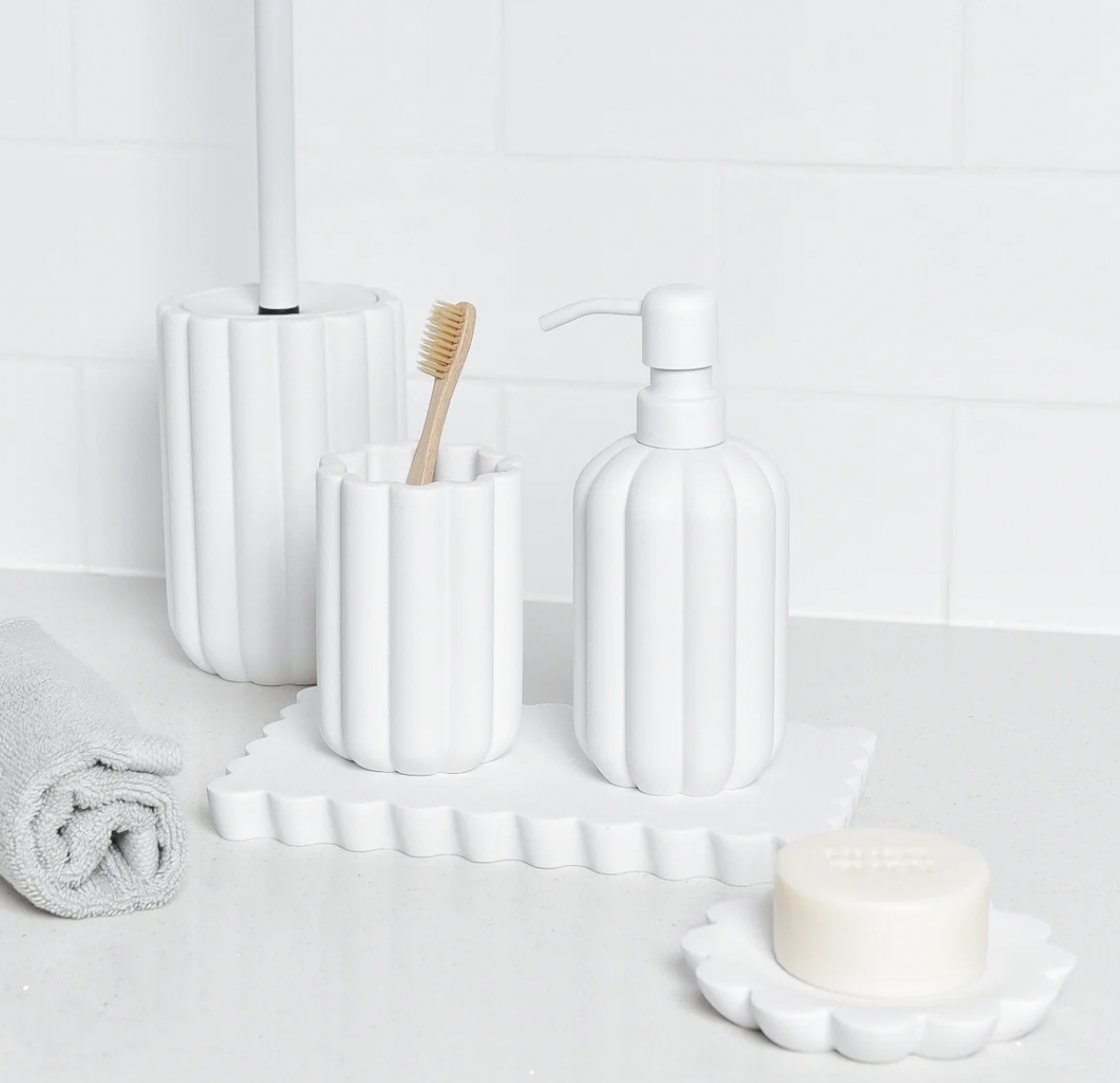 White bathroom accessories including a soap dispenser, toothbrush holder with a toothbrush, tissue box, and rolled towel on a white countertop against a white tiled wall.
