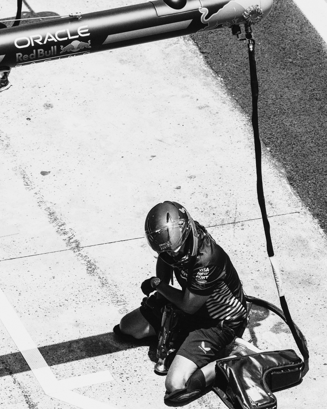 A Forumla 1 Red Bull mechanic kneeling on the ground in the pit lane, wearing a helmet and racing suit, folding his gloves. An arc of a barrier or sign and part of a race car with sponsorship logos are visible overhead.