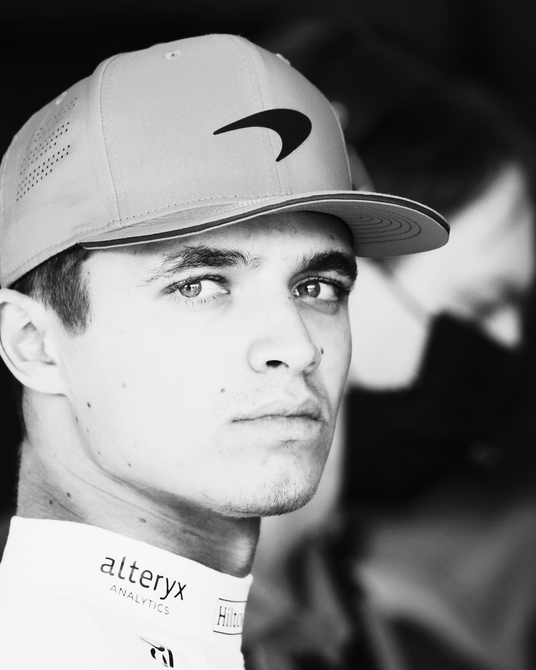 McLaren driver, Lando Norris in a baseball cap and racing suit, looking at the camera, in black and white.