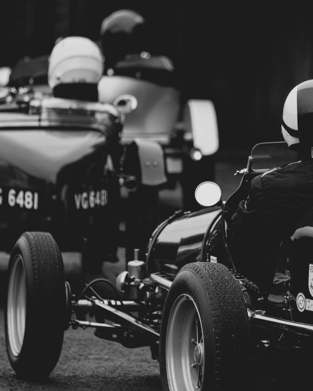 Black and white photo of vintage race cars with drivers wearing helmets, parked closely together.