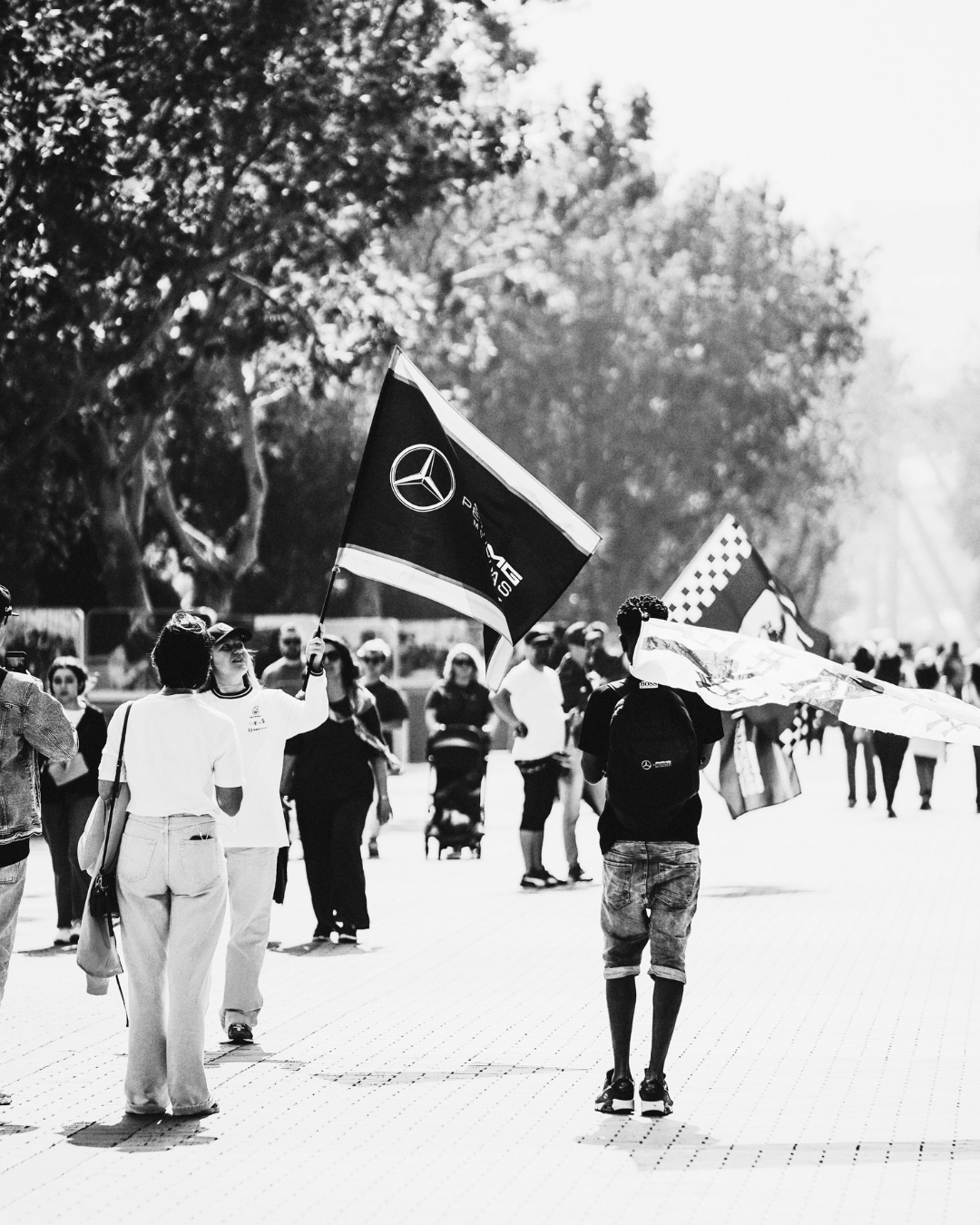People walking with flags in an outdoor event on a sunny day, some holding Mercedes-Benz flags, surrounded by trees.