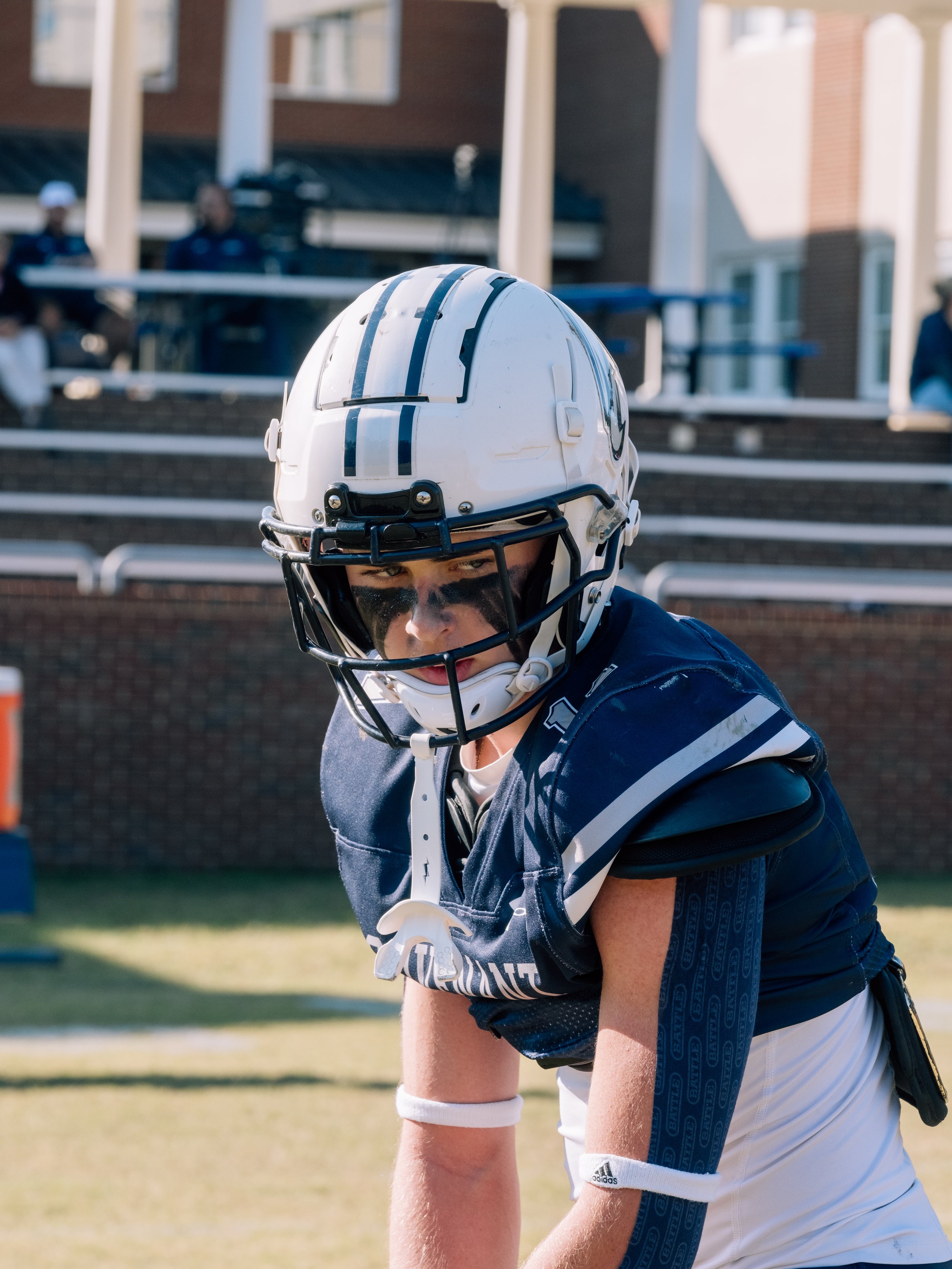A young football player wearing a white helmet with the number 24, black eye paint, a navy blue jersey with white accents, on a football field.