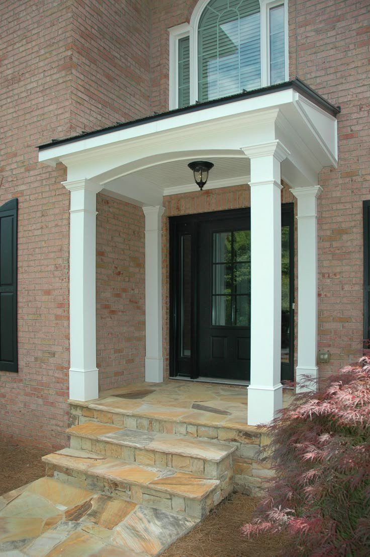 Front porch of a brick house with stone steps, white columns, a black door, and a small balcony with a window above.