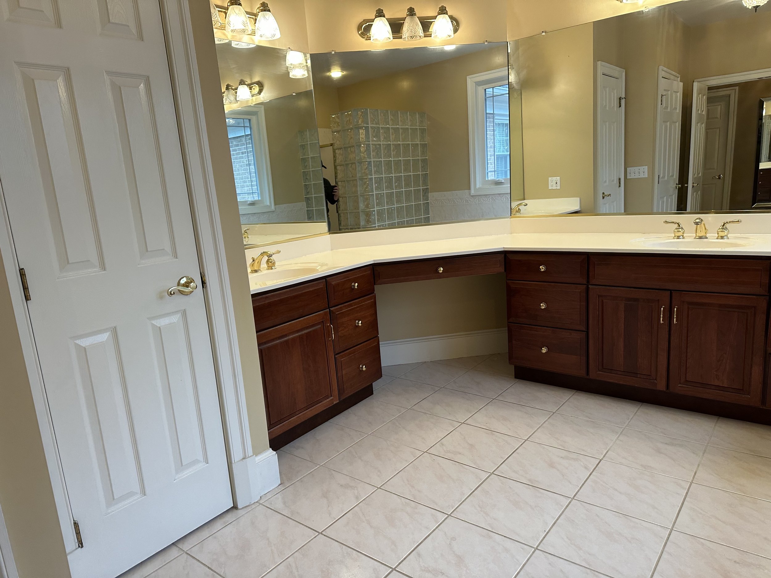 A bathroom vanity with a large mirror, gold fixtures, wooden cabinets, beige tiled floor, and a window reflection in the mirror.