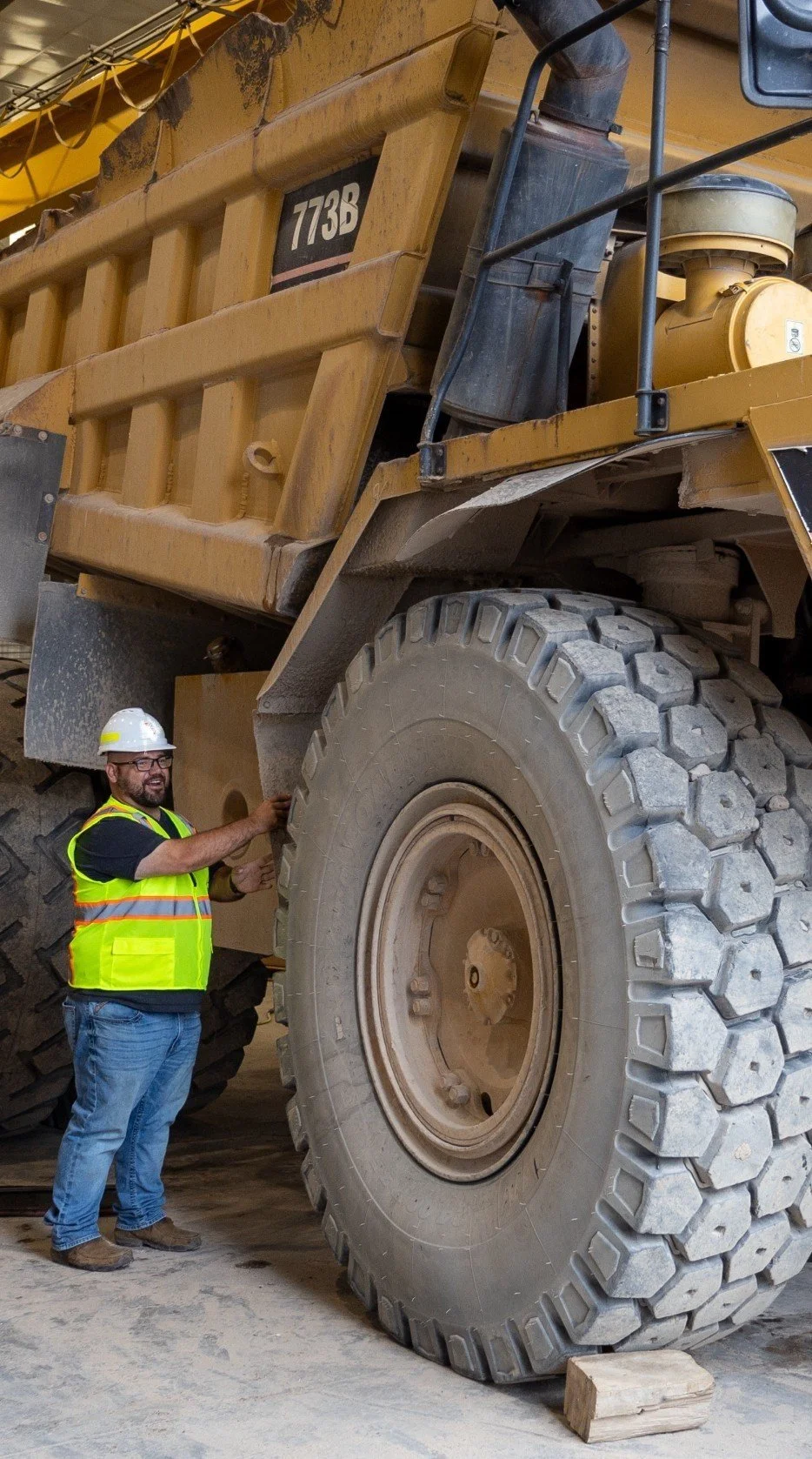 A construction worker wearing a white safety helmet and yellow vest examining the large tire of a yellow dump truck in a construction site.