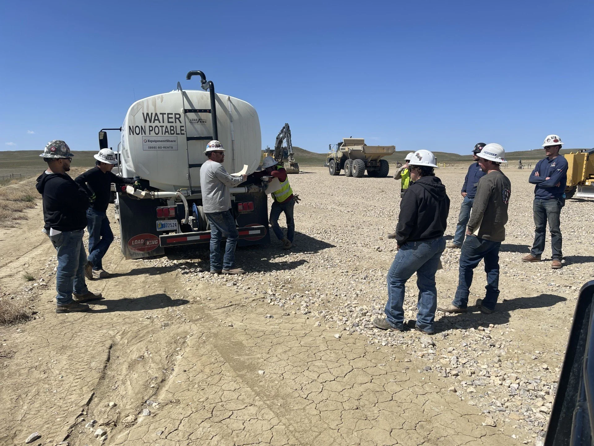 Group of workers and engineers wearing hard hats and safety gear, gathered around a water tanker labeled 'Water Non Potable' on a dry, cracked terrain with construction vehicles in the background under a clear blue sky.
