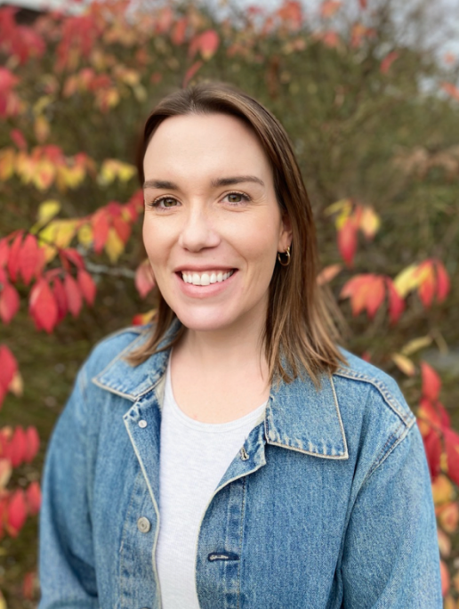A woman with brown hair smiling outdoors in front of a bush with red and yellow leaves, wearing a denim jacket and a white shirt.