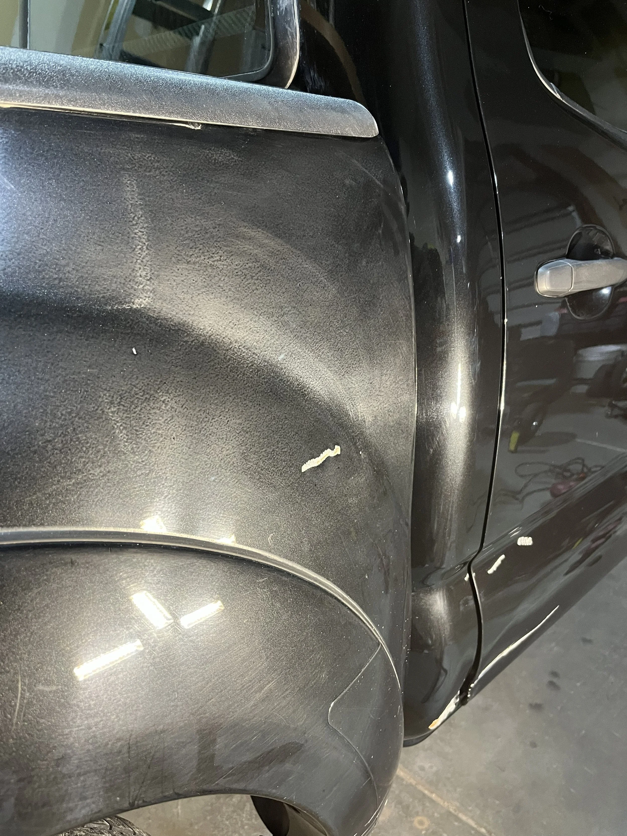 Close-up of a black car's front fender and door, showing minor scratches, a small chip in the paint, and a reflection of the surrounding garage area.