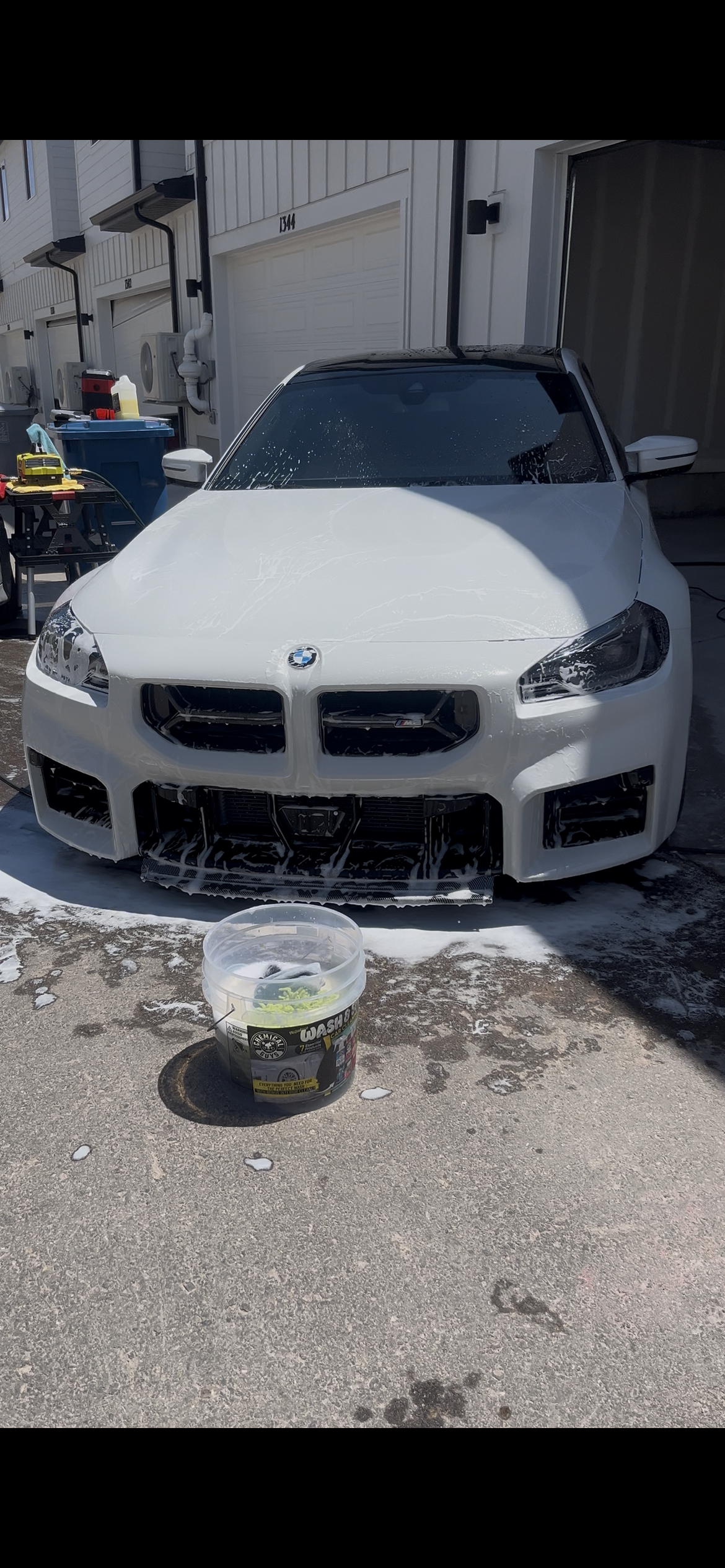 A white BMW sports car being washed with soap on the driveway of a residential area, with cleaning supplies and tools nearby.