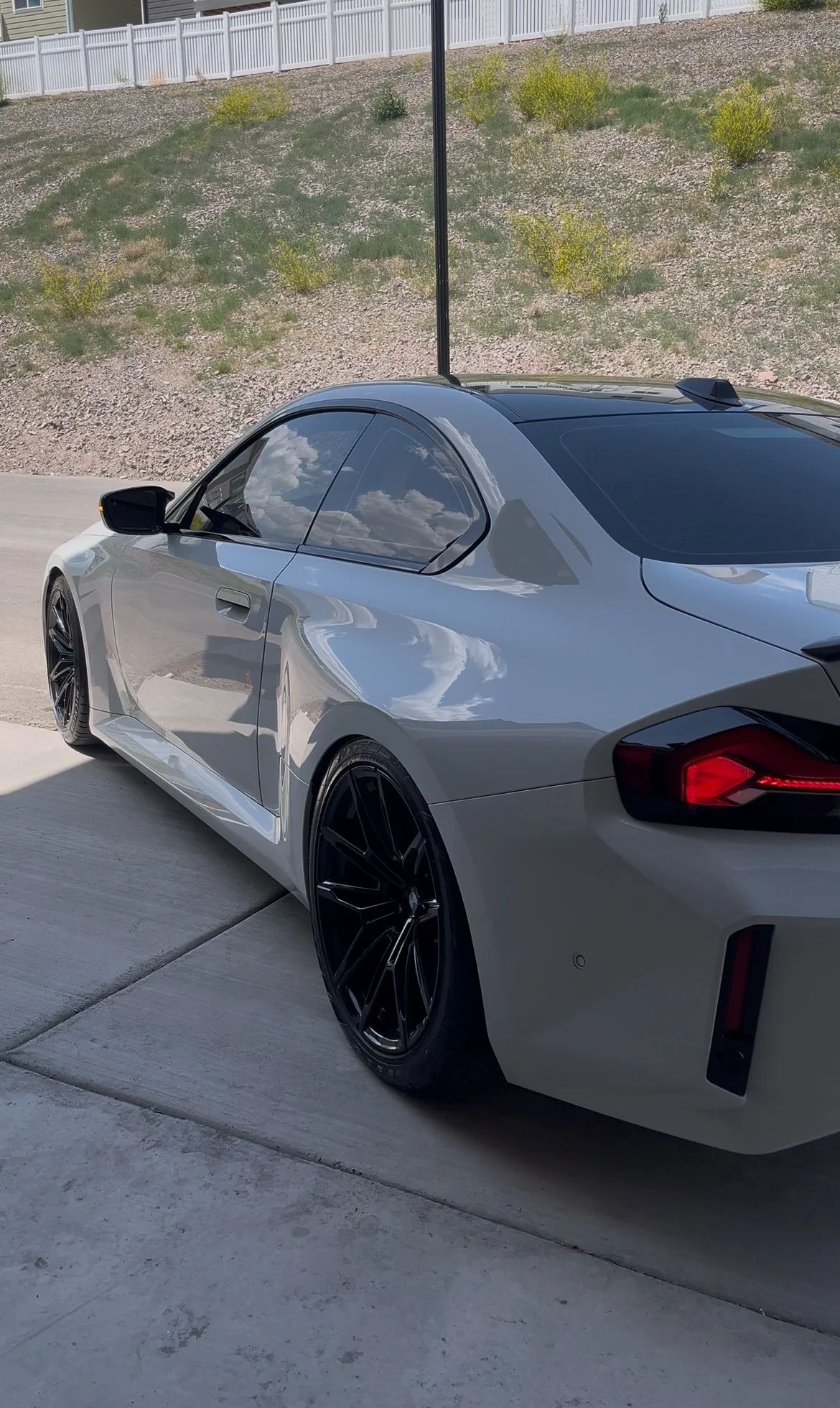 Silver sports car with black wheels parked on a concrete driveway, reflecting the sky and clouds in its windows, with a white fence and dirt hillside in the background.
