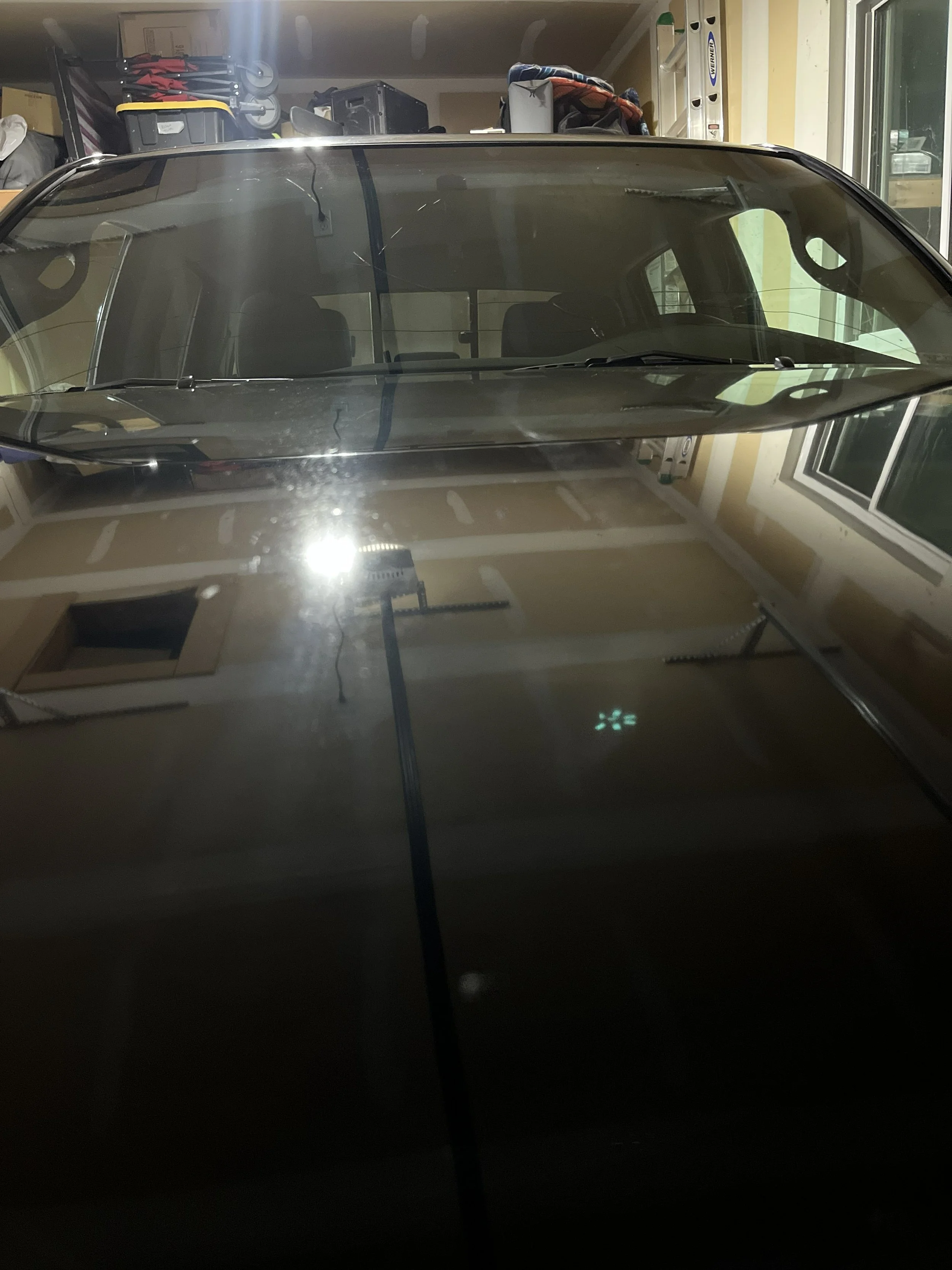 A black car is parked in a garage with a glass windshield reflecting the light. The garage contains various items on shelves, including storage bins, boxes, and a level tool.
