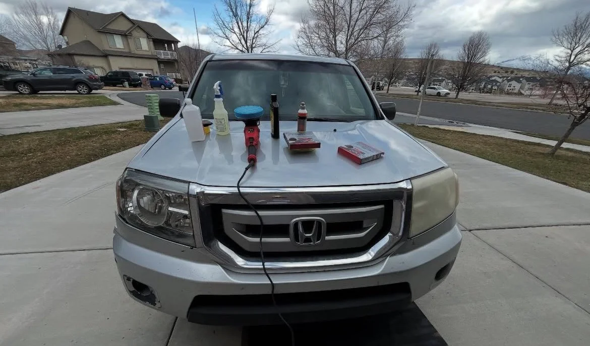 Front view of a silver Honda SUV parked on a driveway with various car cleaning supplies and tools placed on the hood, including a spray bottle, cleaning cloths, and polishing or detailing tools.