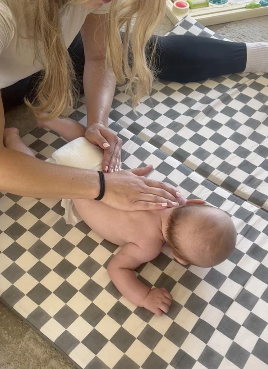 Close-up of a therapist's hands supporting a baby through tummy time to build motor strength, in-home visit in Costa Mesa, CA.