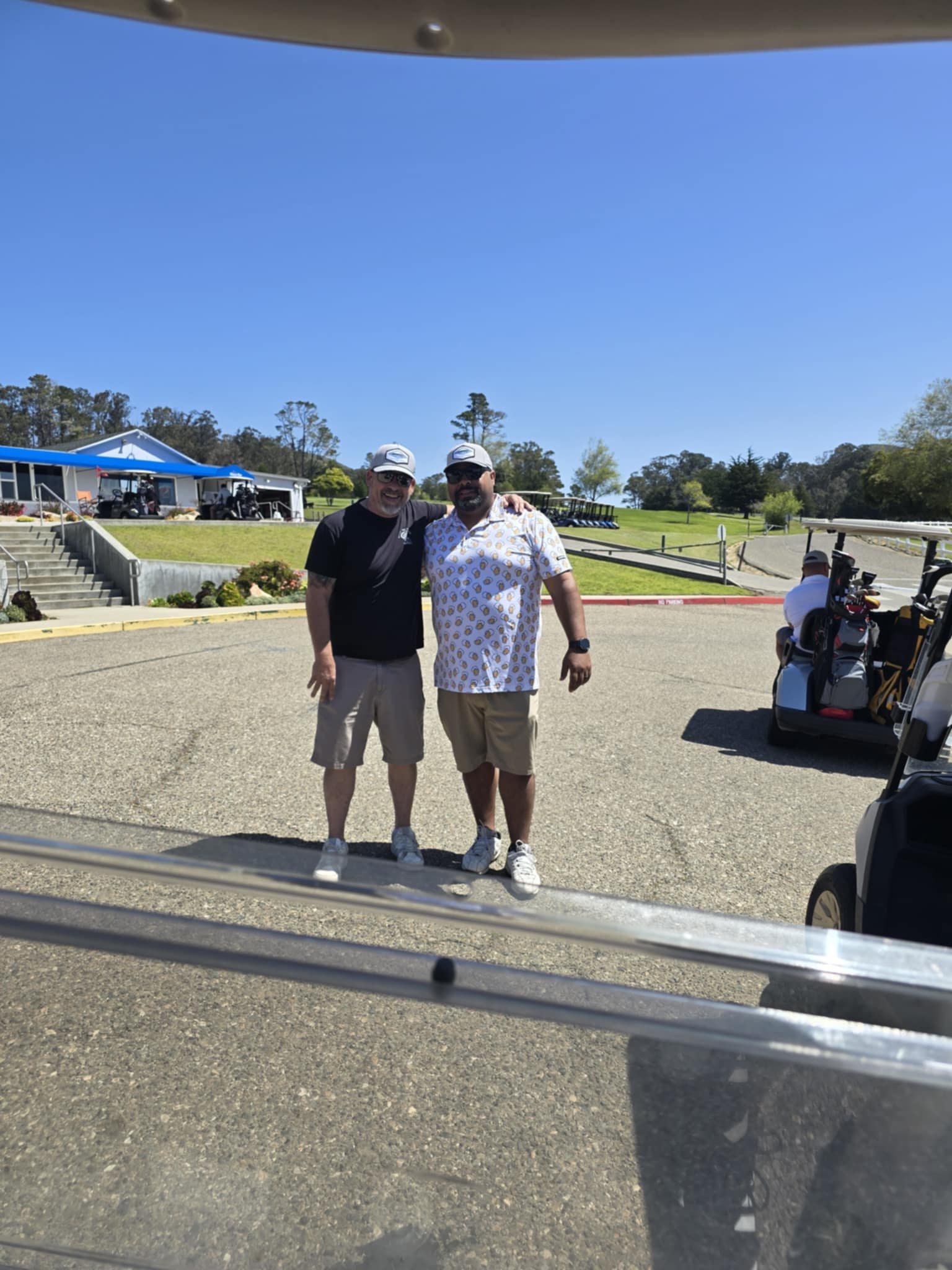 Two men standing arm in arm on a golf course with golf carts and clubhouse in the background, on a sunny day.