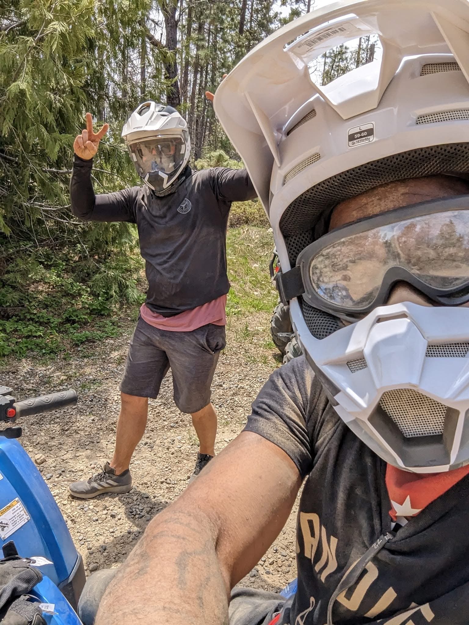 Two people wearing helmets and goggles taking a selfie on a dirt trail surrounded by trees, one making a peace sign.