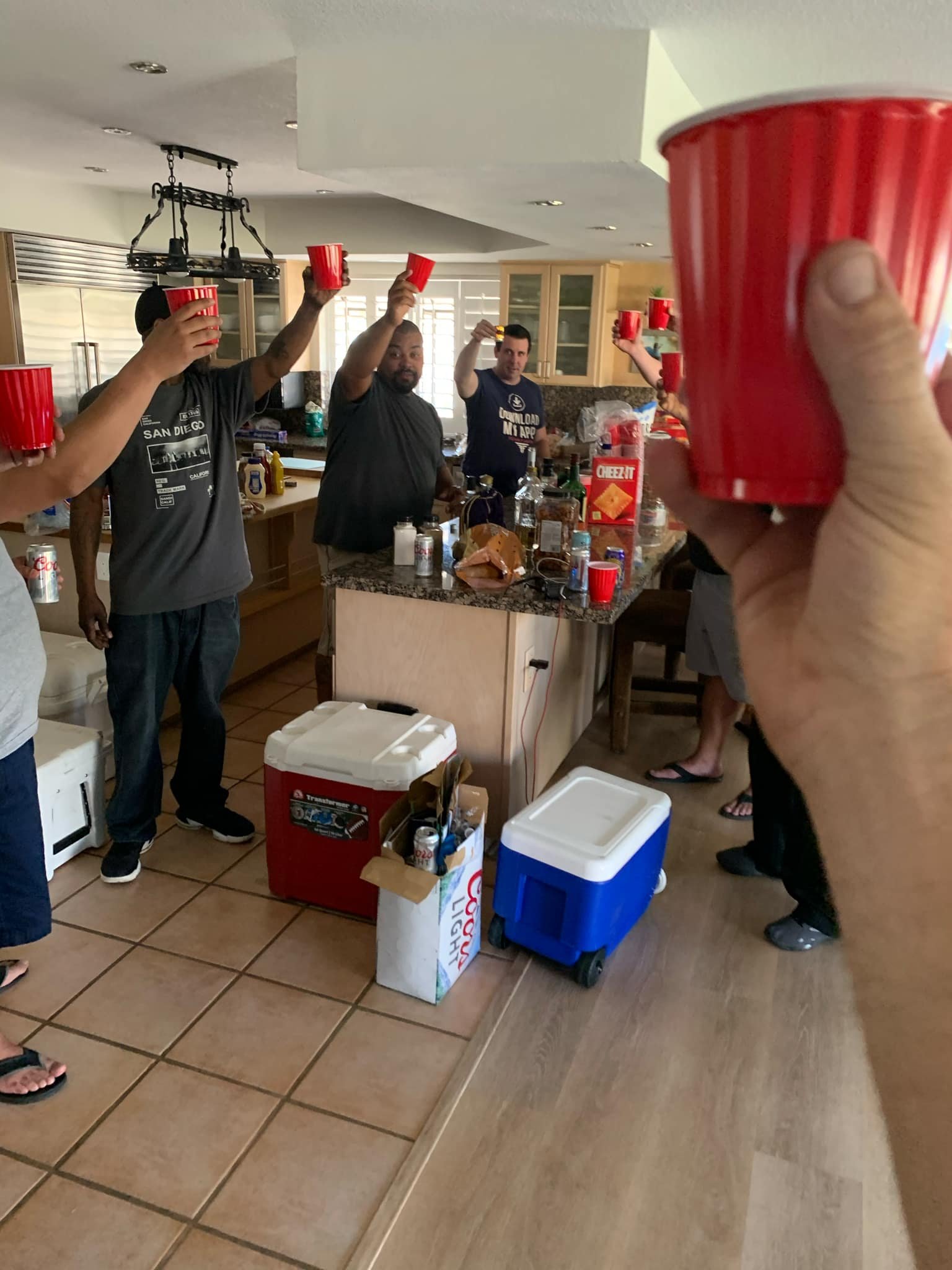 People raising red cups in a toast in a kitchen with a group of friends, celebrating around a counter with food and drinks.