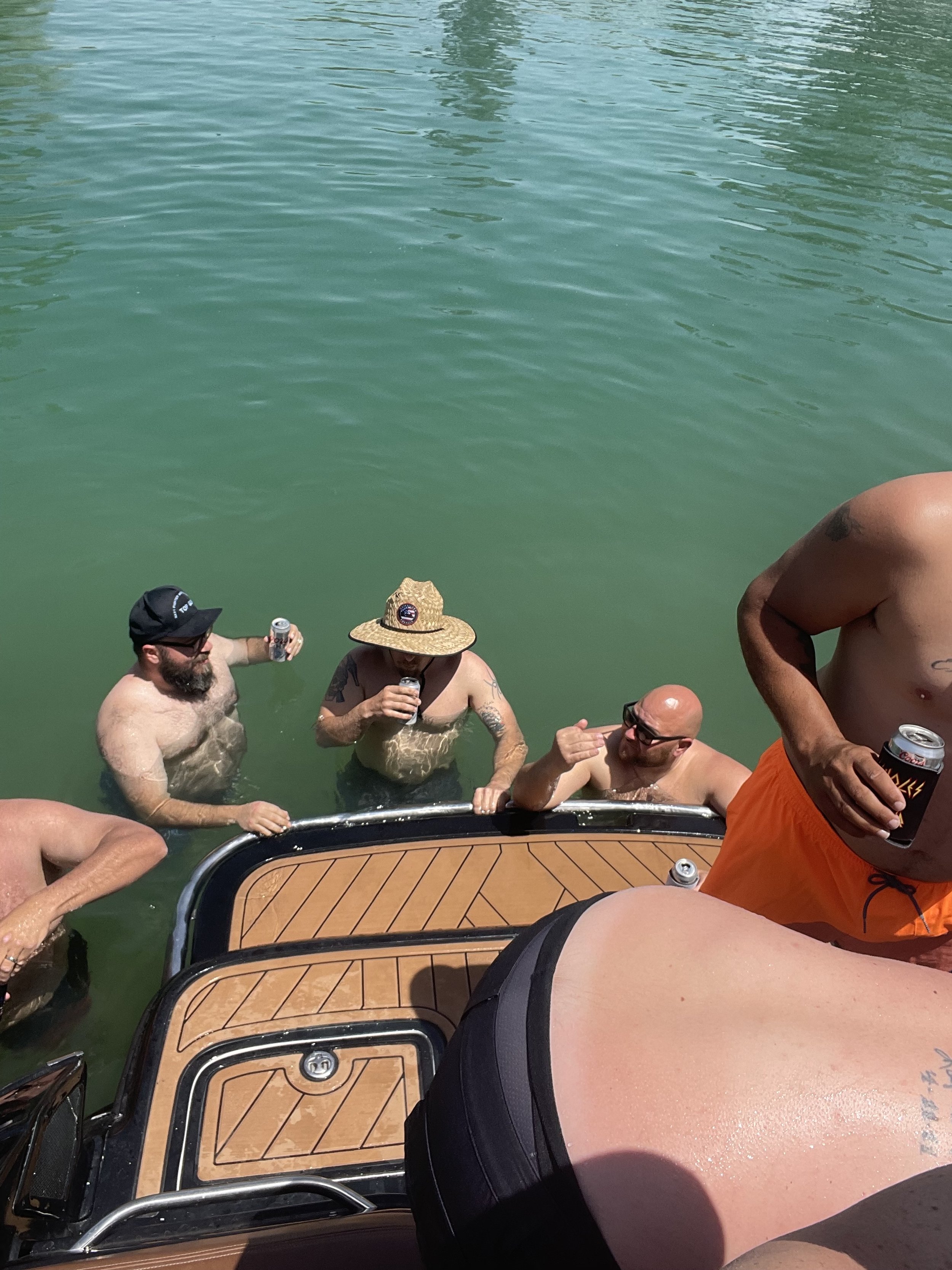 Group of men in swimwear and sunglasses socializing on a boat in a body of water, some holding cans or drinks.
